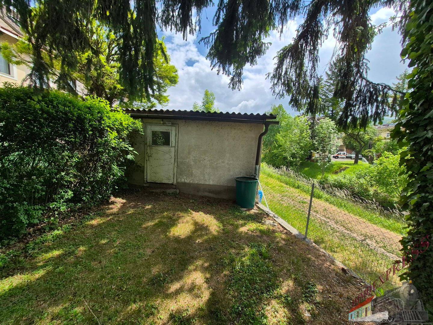 Kleiner Gartenschuppen am Hang mit einfacher Tür und Mülltonne im Grünen.