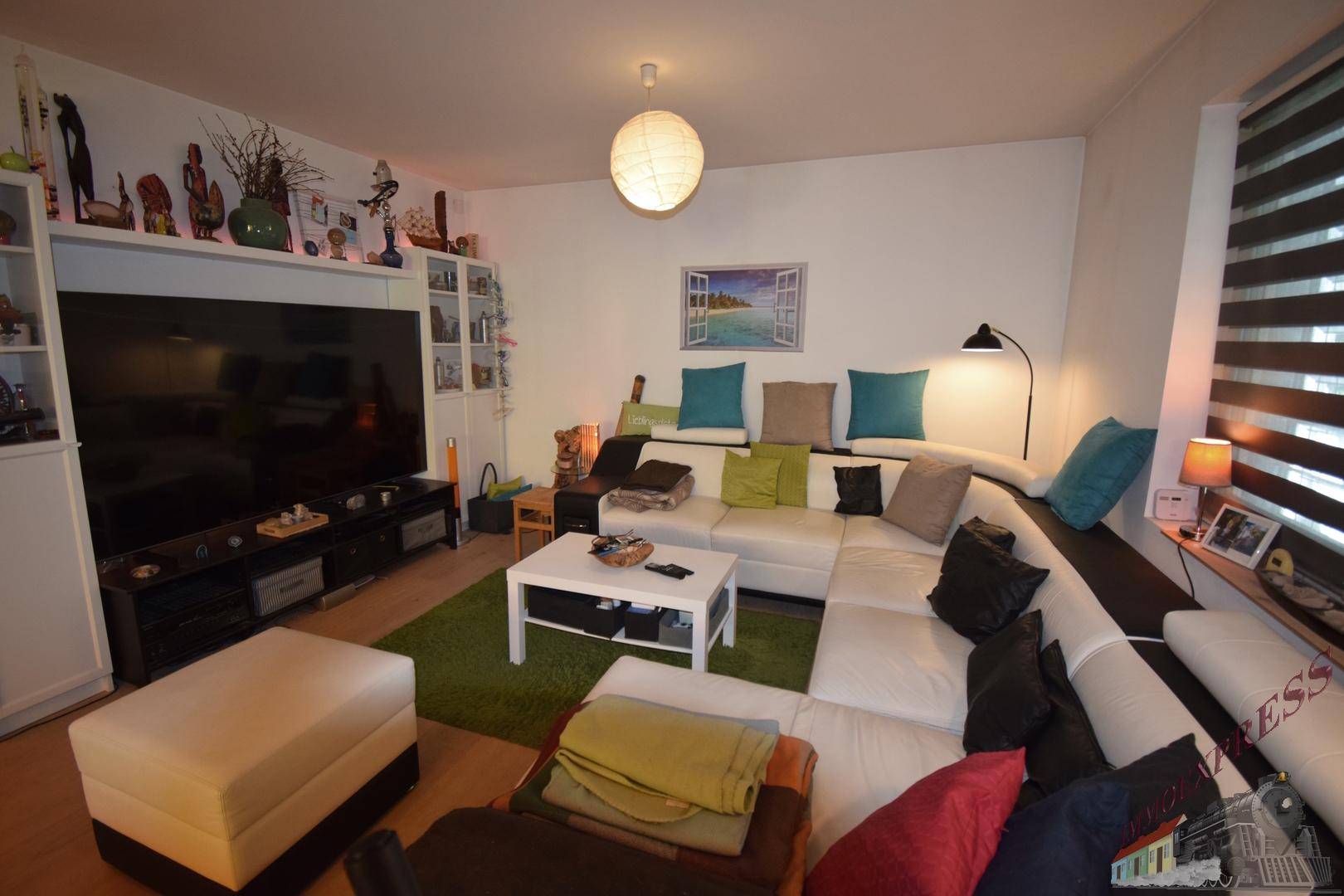 Gemütliches Wohnzimmer mit großer weißer Eckcouch, Fernseher und farbigen Kissen.