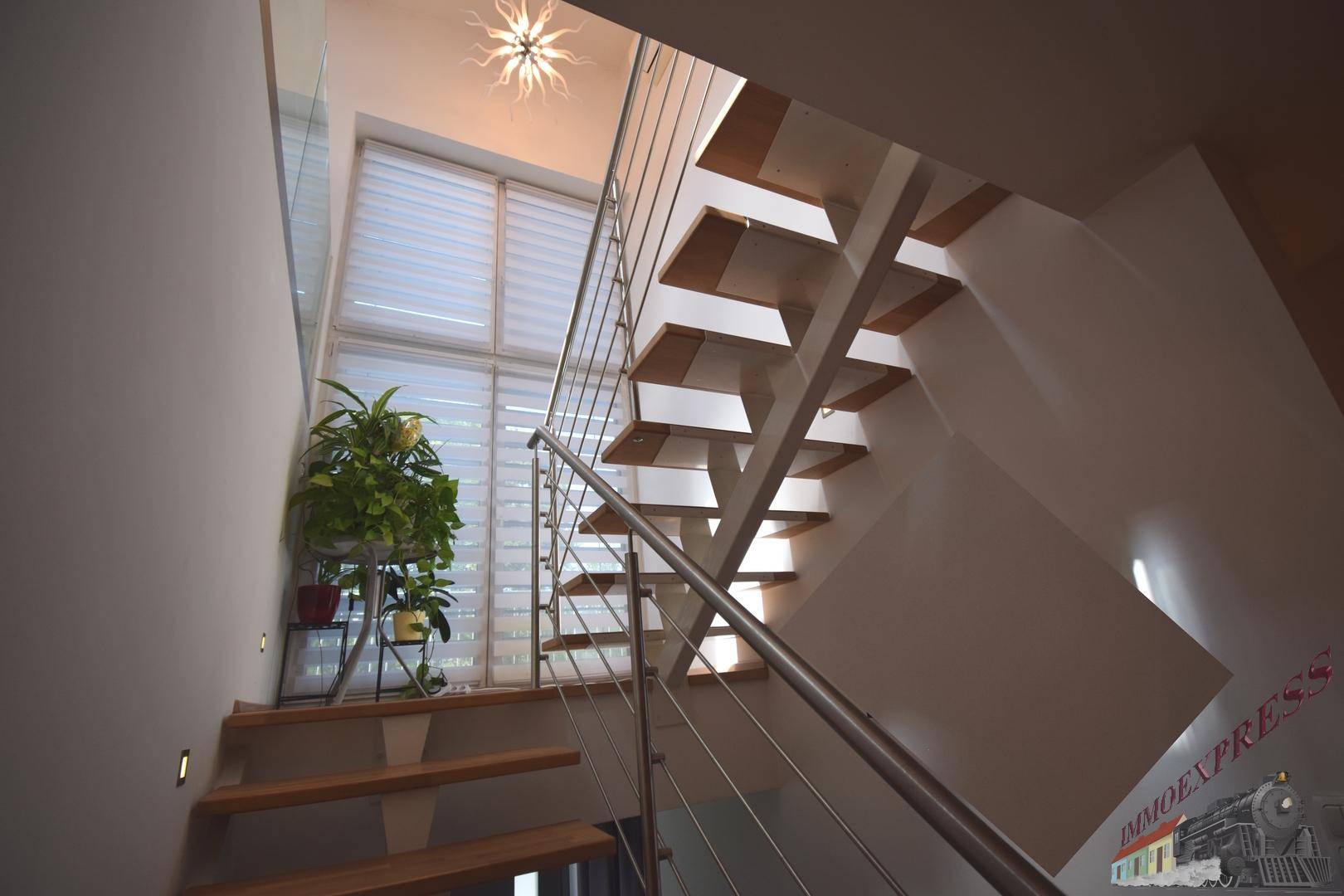 Moderne Holztreppe mit Edelstahlgeländer und großen Fenstern, die viel Licht hereinlassen.