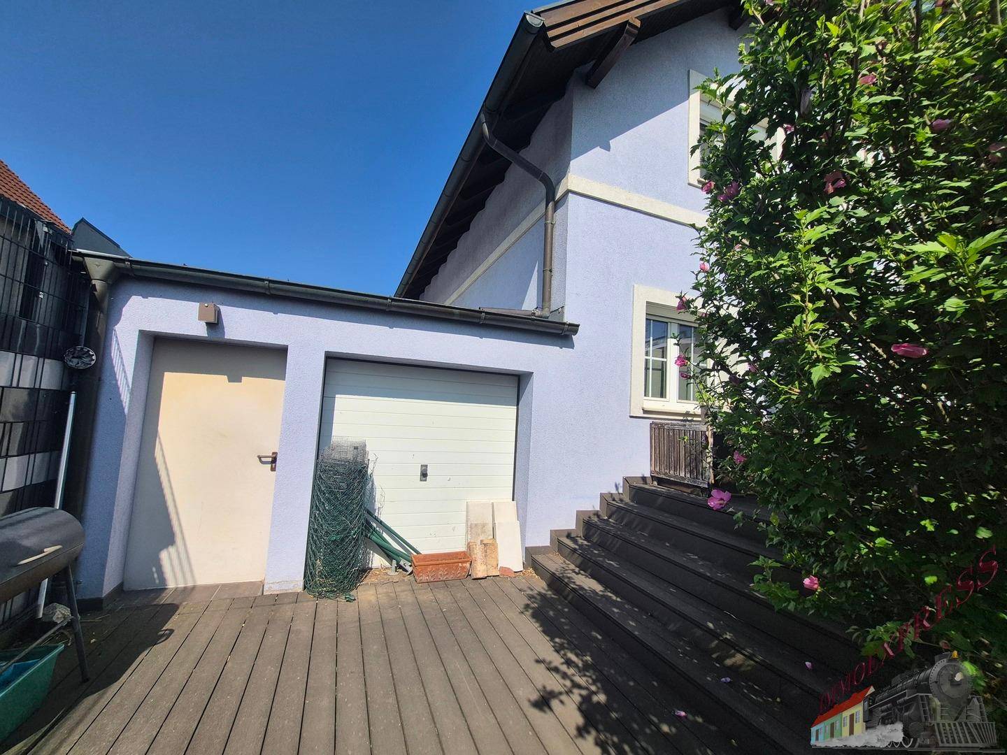 Seitlicher Anbau mit Garage und einer Holzterrasse, die zu einem Eingang führt.