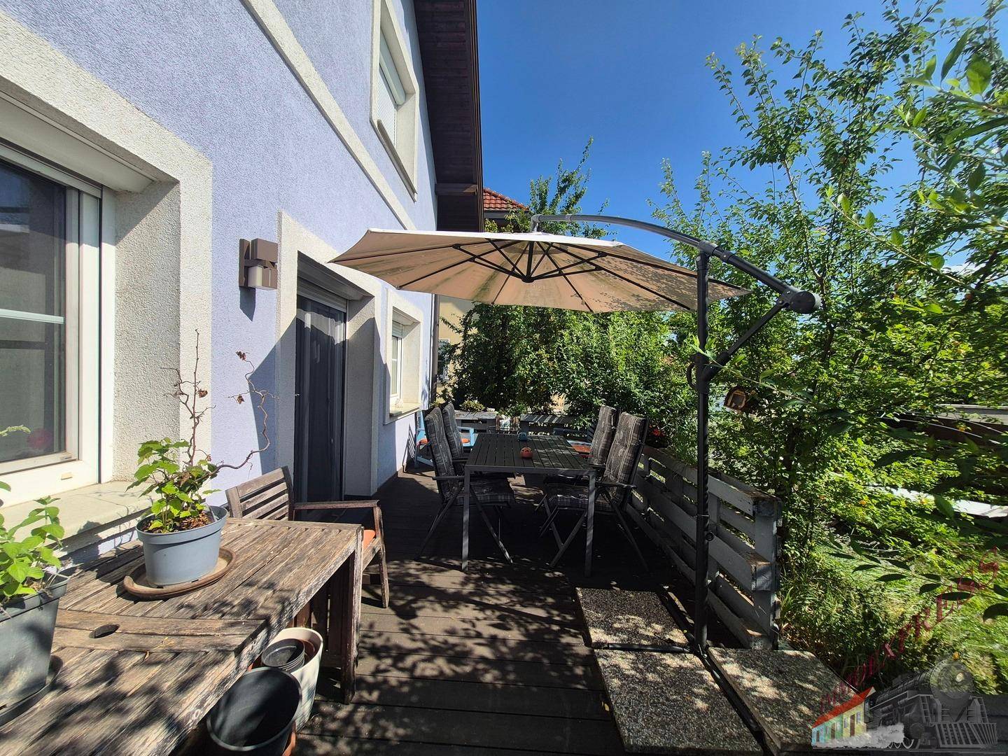 Gemütliche Holzterrasse mit Esstisch, Stühlen und Sonnenschirm, umgeben von viel Grün.