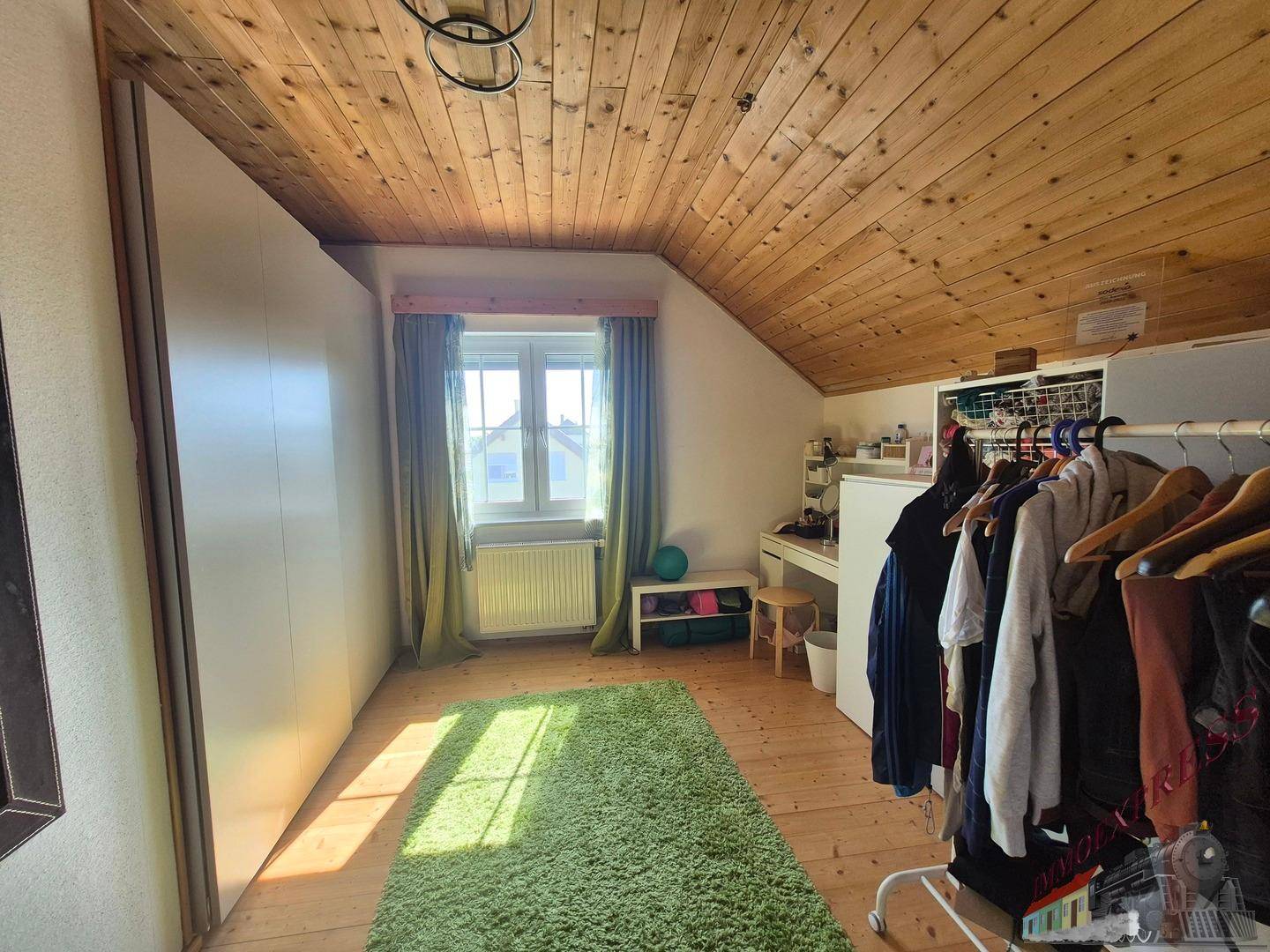 Schlafzimmer im Dachgeschoss mit Holzboden, grüner Teppich und Kleiderstange.