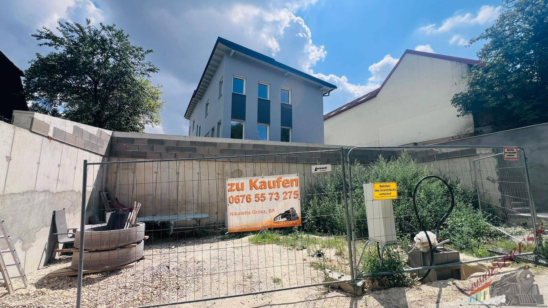 Neubau-Einfamilienhaus von der Straße aus gesehen, umgeben von einem Bauzaun und einem "Zu Kaufen"-Schild.