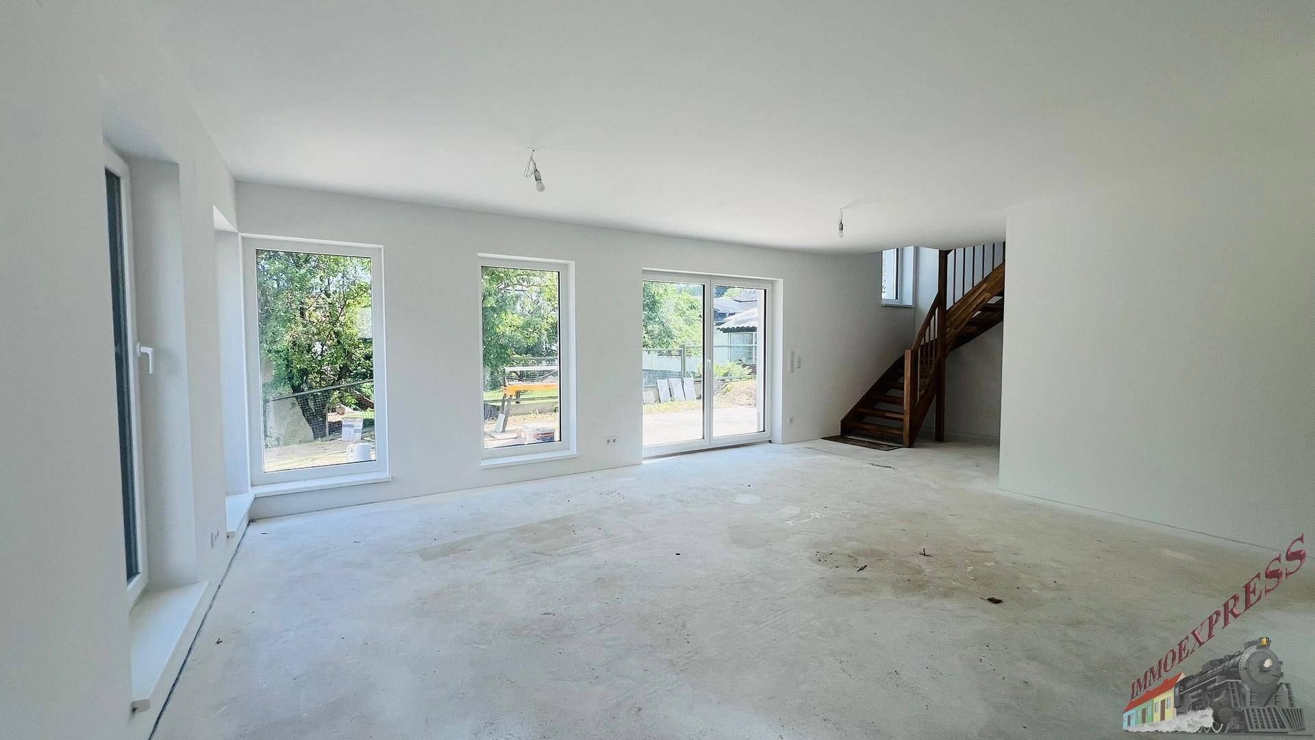 Geräumiger Wohnbereich im Neubau mit Betonboden und einer Holztreppe, die in die oberen Stockwerke führt.