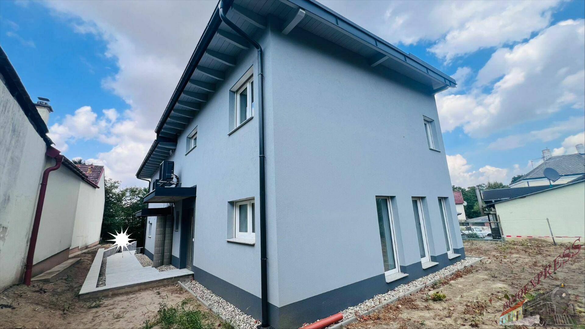 Seitenansicht des modernen Neubaus mit heller Fassade, dunklem Sockel und überdachtem Eingangsbereich.
