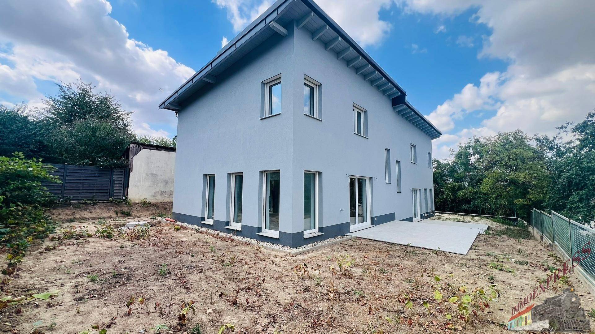Rückansicht des Neubaus mit großer Terrasse und dem noch ungestalteten Gartenbereich unter blauem Himmel.