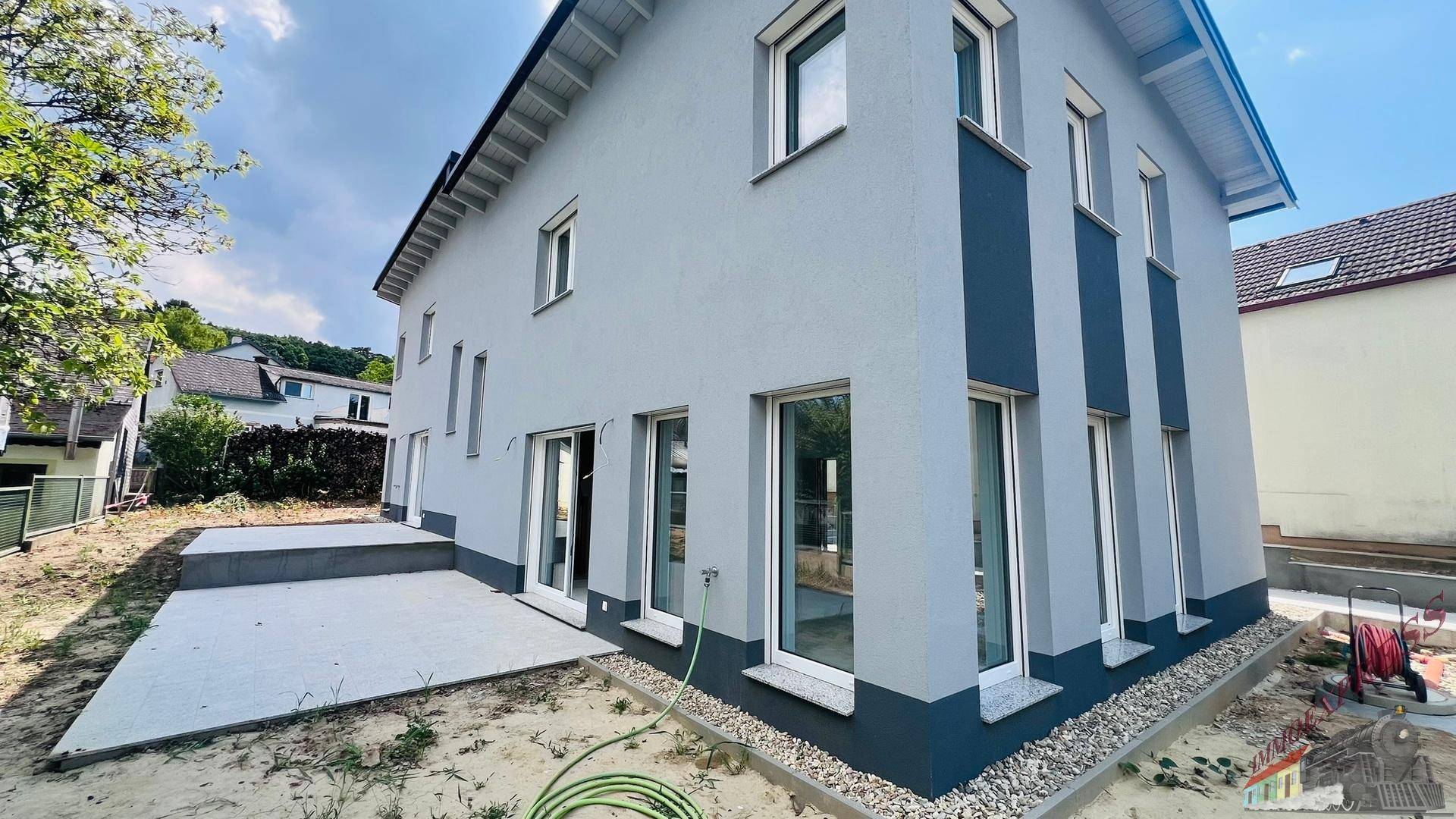 Seitenansicht des Neubaus mit moderner Fassadengestaltung, großen Fenstern und einer betonierten Terrasse.