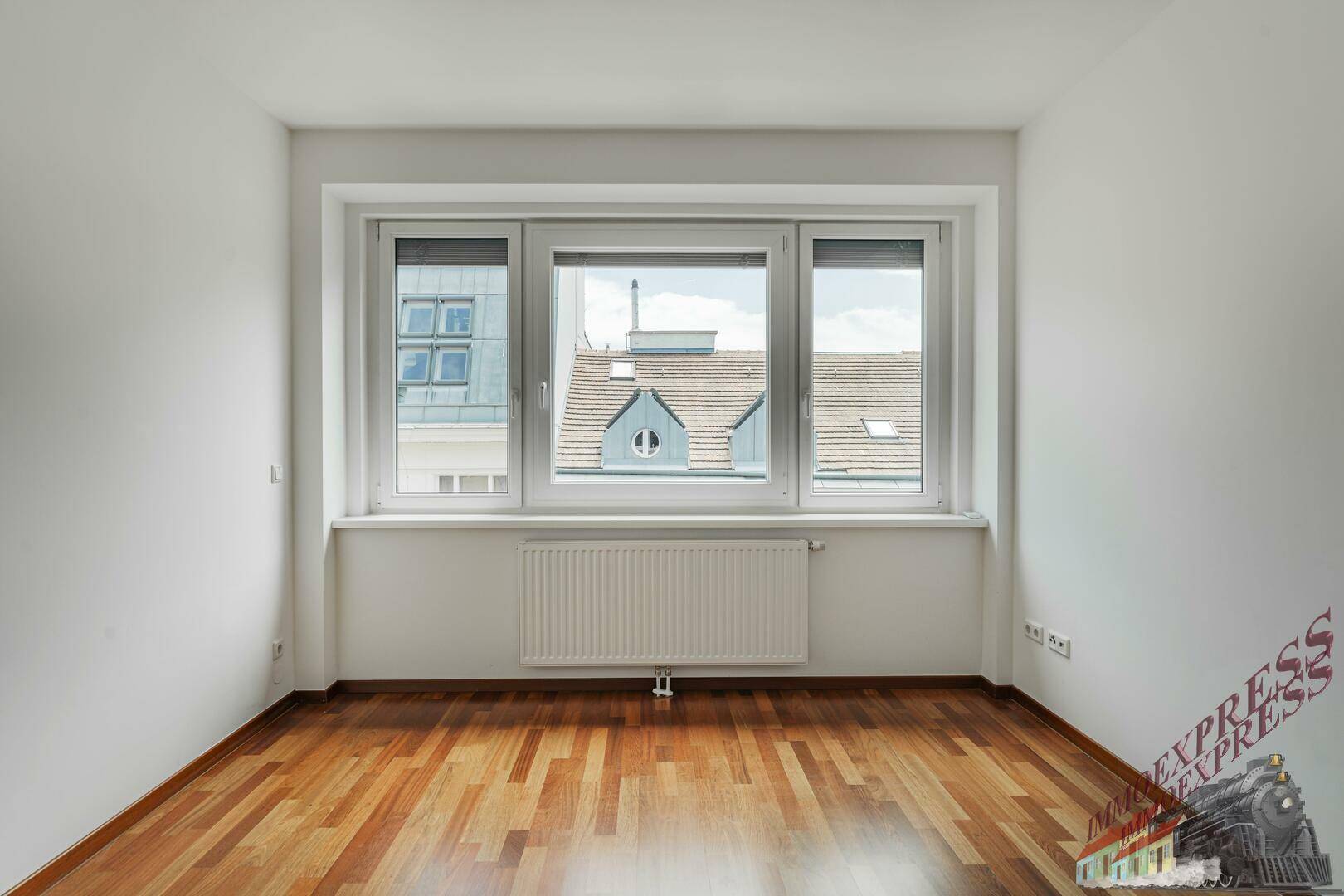 Helles Zimmer mit Parkettboden, großen Fenstern und Blick auf die Dächer.