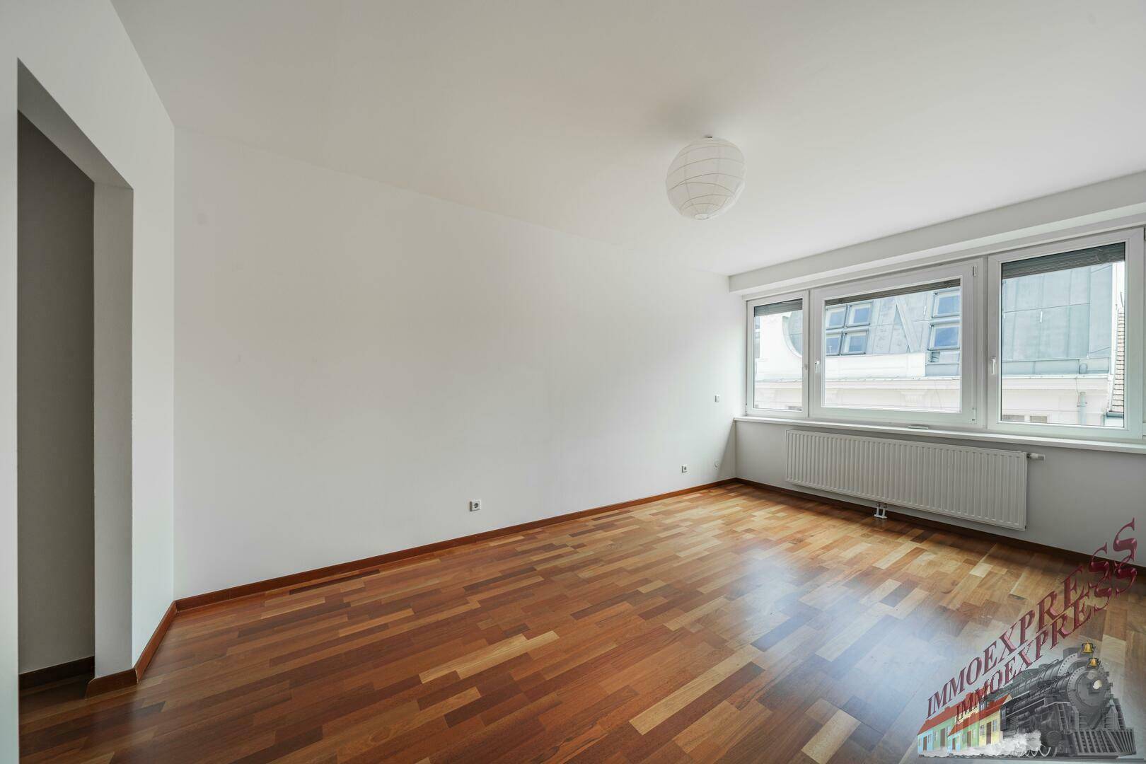 Helles Zimmer mit Parkettboden, großen Fenstern und Blick auf die Dächer.