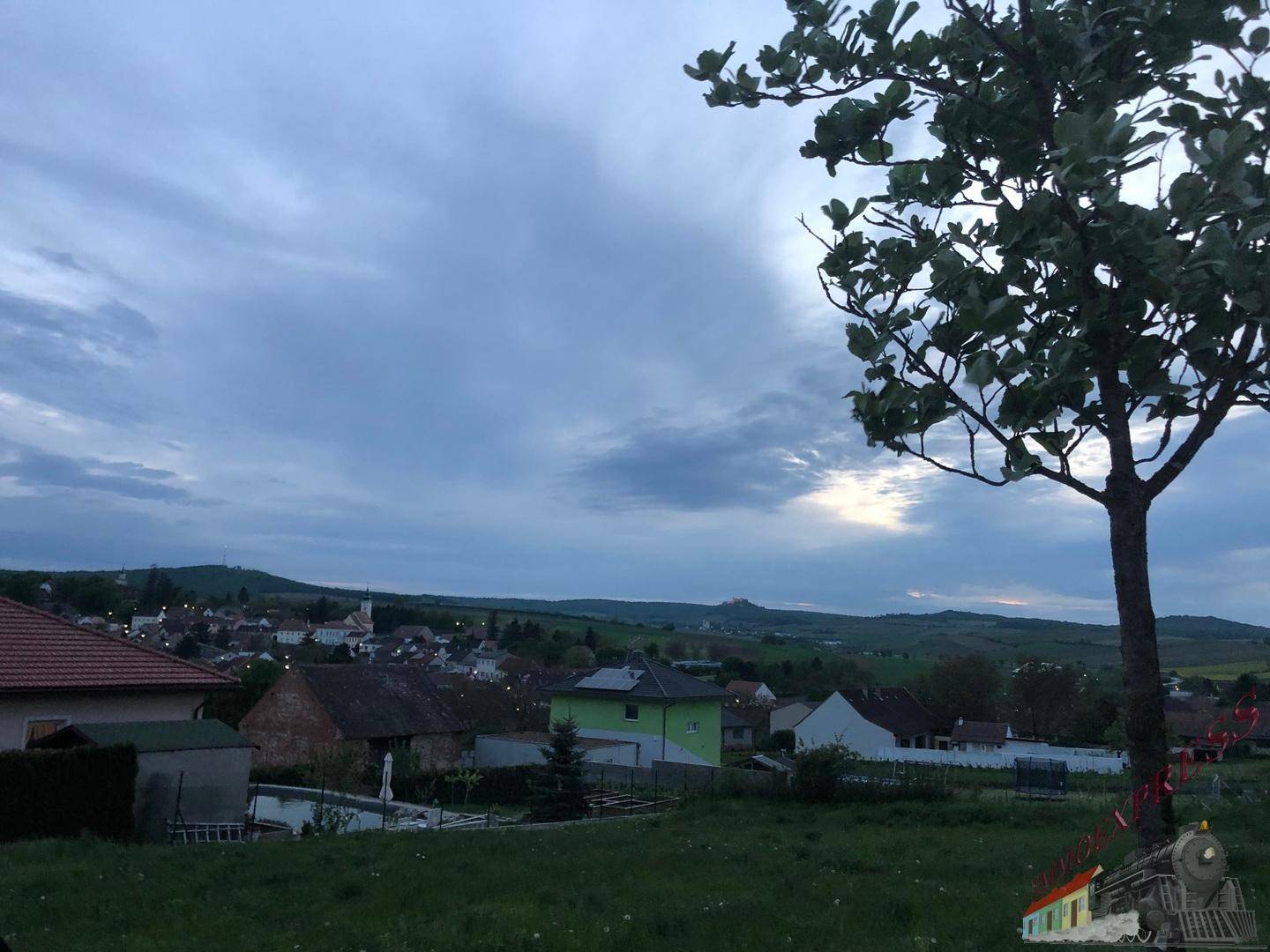 Abendlicher Blick über die Landschaft mit Häusern und einem Baum im Vordergrund unter bewölktem Himmel.