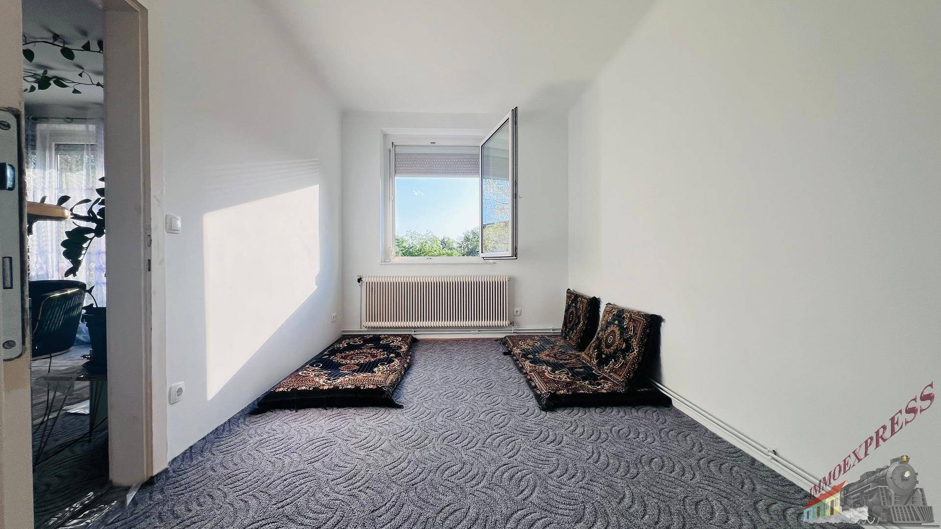 Helles Zimmer mit Teppichboden, Fenster und Heizkörper, derzeit mit Bodenkissen eingerichtet.