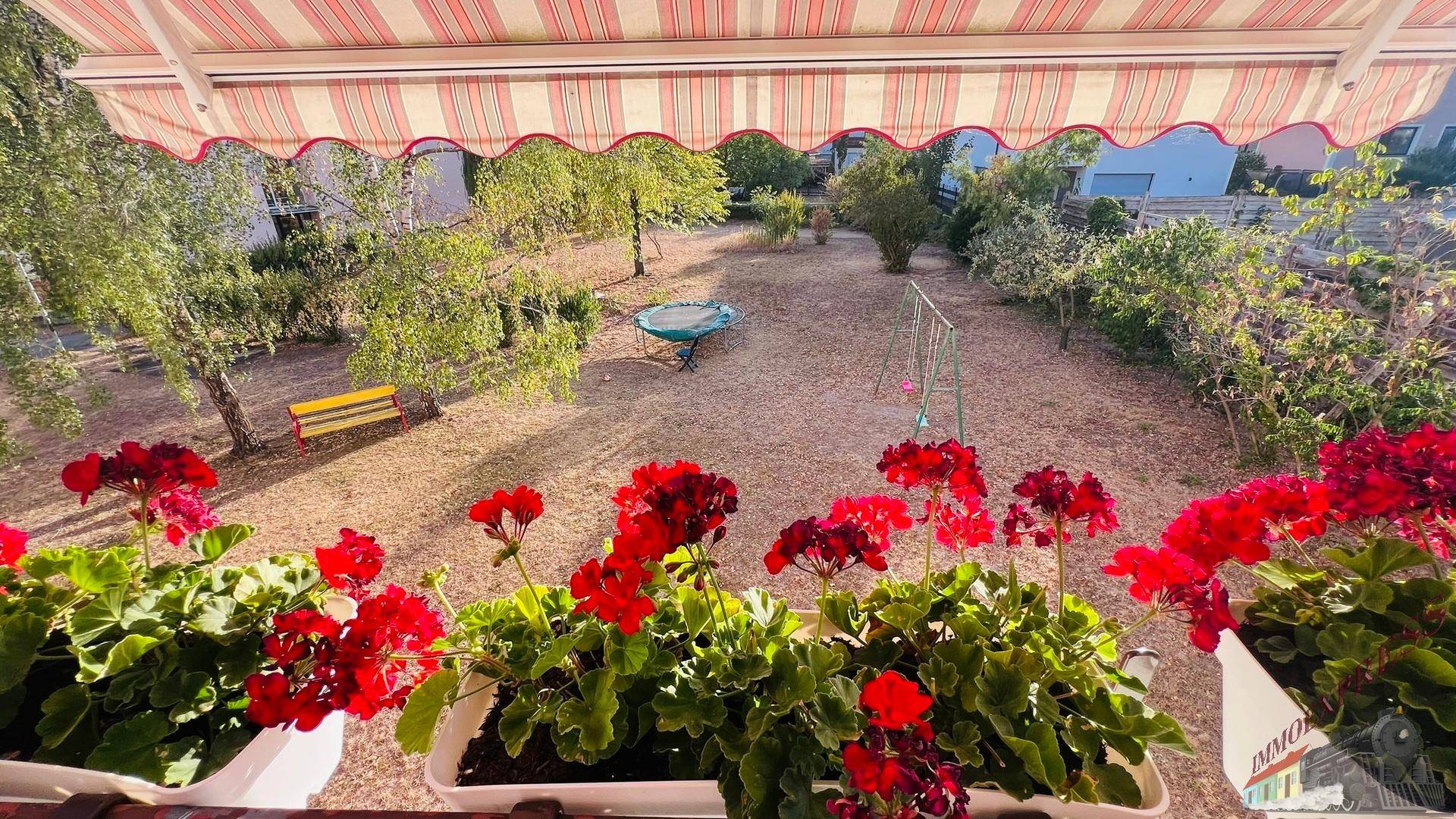 Blick vom Balkon auf den Garten mit Schaukel und Bäumen, rote Blumen im Vordergrund.