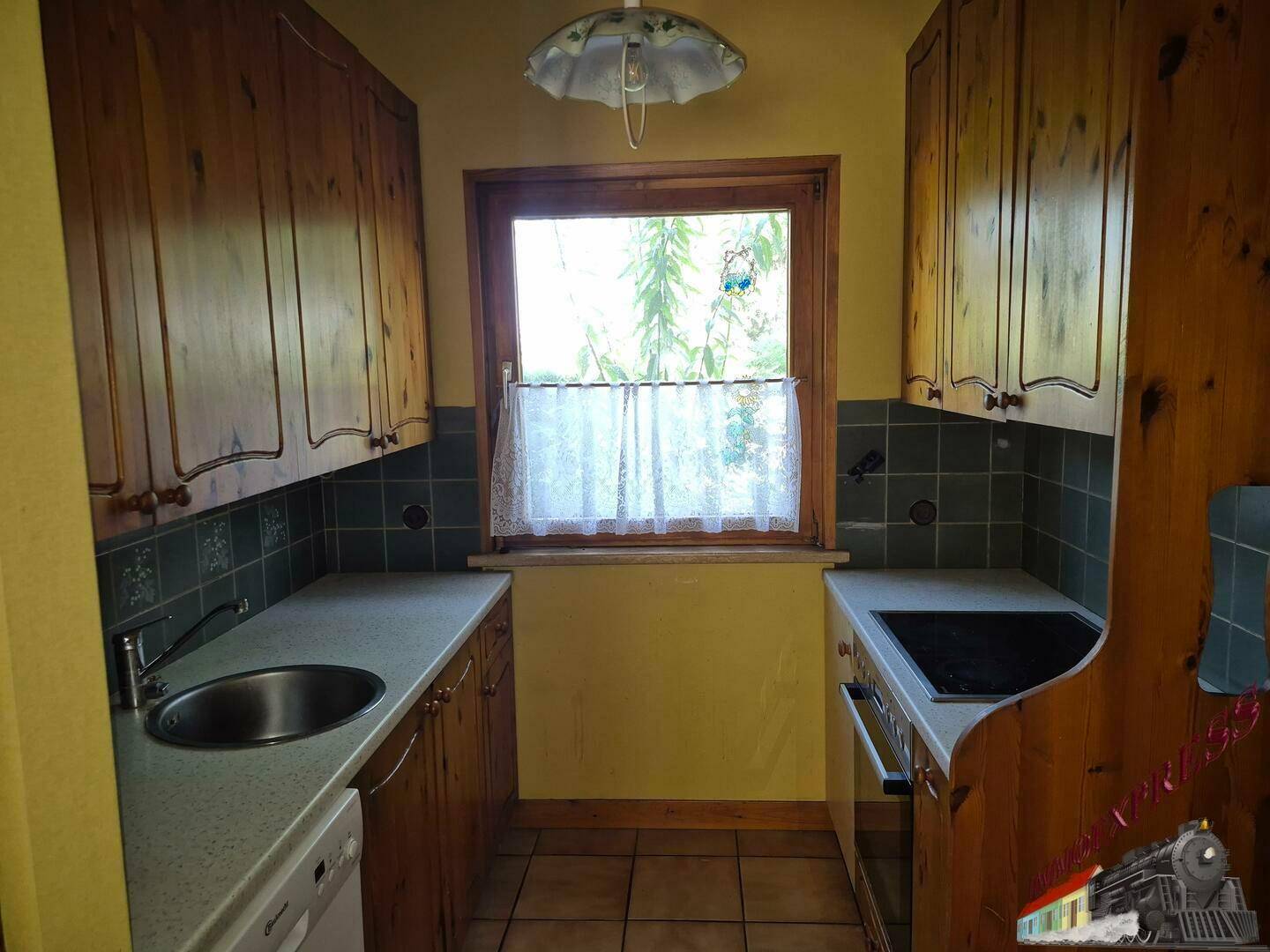 Kleine Küche mit Holzschränken, Spüle, Kochfeld und Fenster mit Gardine.