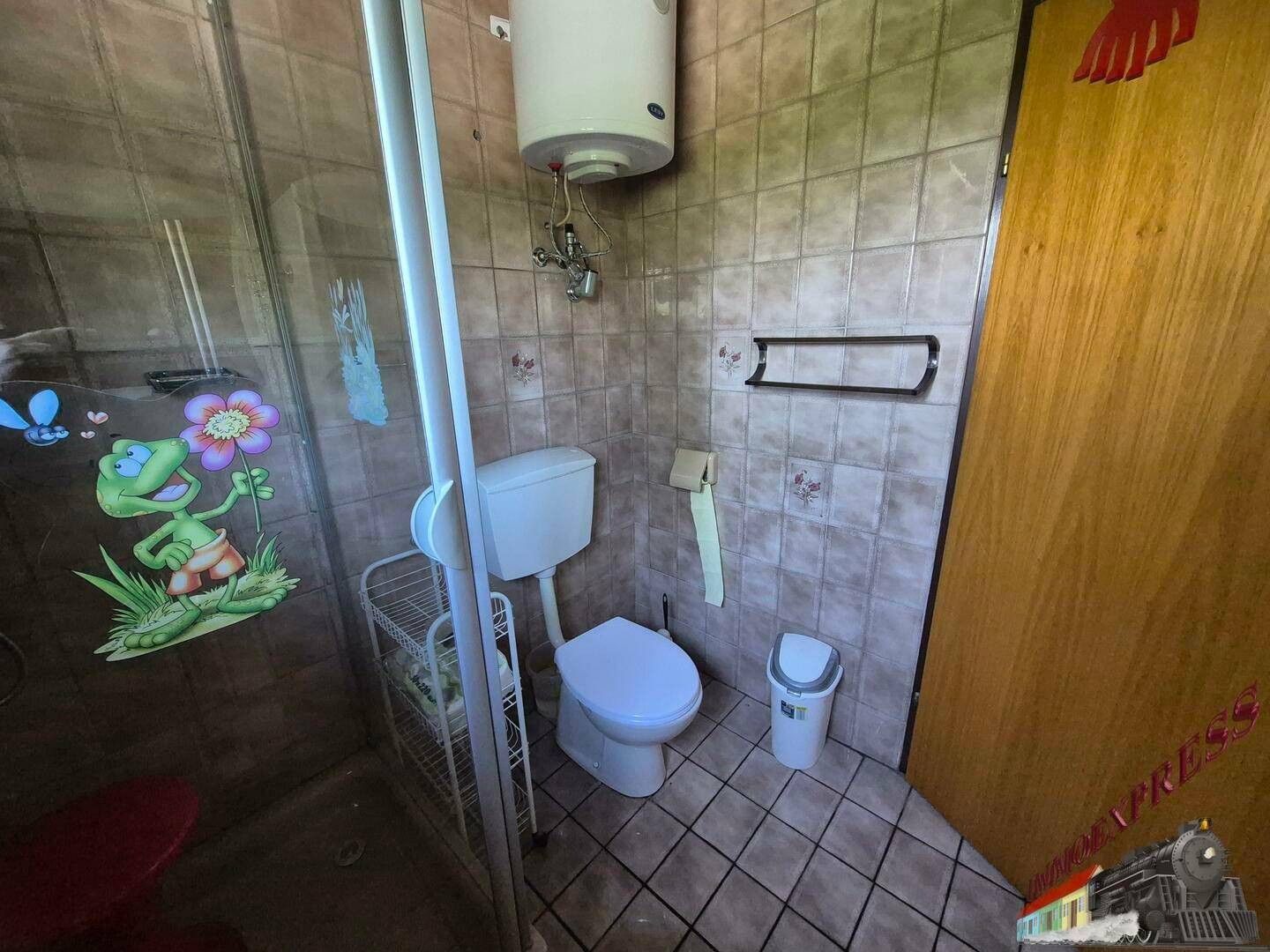 Kleines WC mit Dusche, Toilette und Warmwasserboiler, gefliest in Brauntönen.