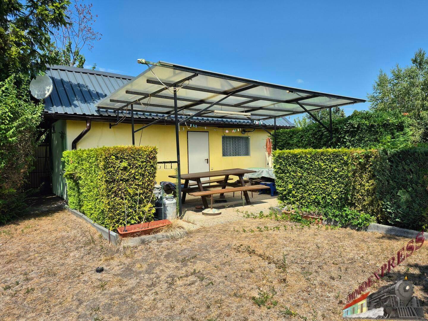 Kleines Haus mit überdachter Terrasse und Gartenmöbeln, umgeben von Hecken und trockenem Rasen.