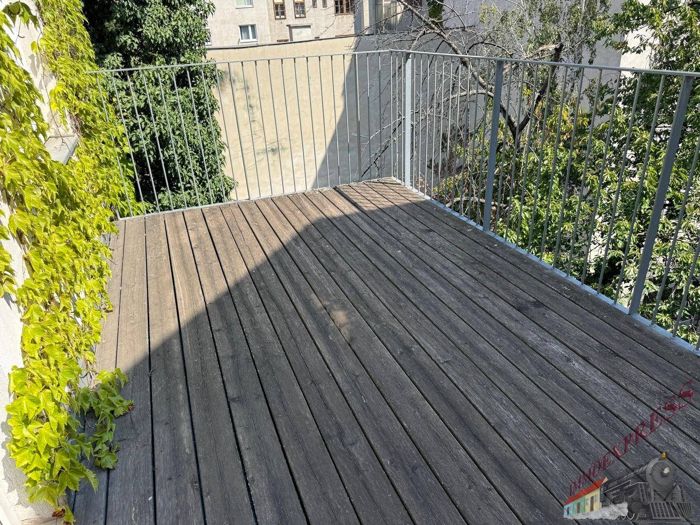 Sonniger Balkon mit Holzboden und Metallgeländer, umgeben von städtischer Vegetation.