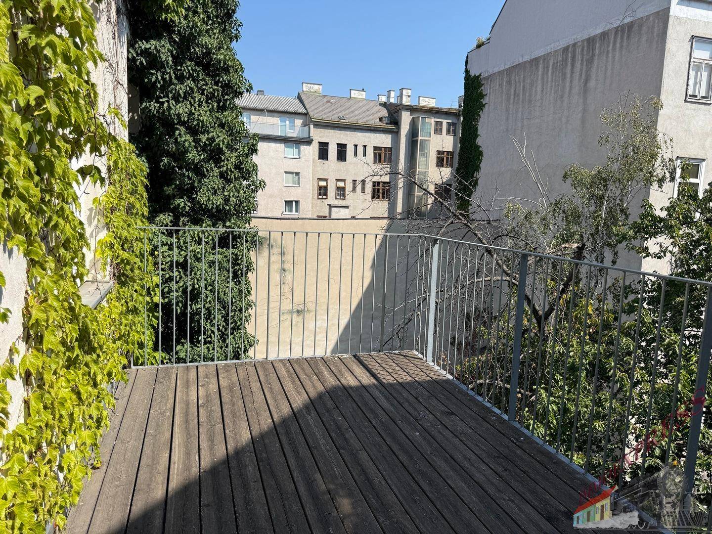 Balkon mit Holzbelag und Blick auf die umliegenden Gebäude und grüne Bäume.