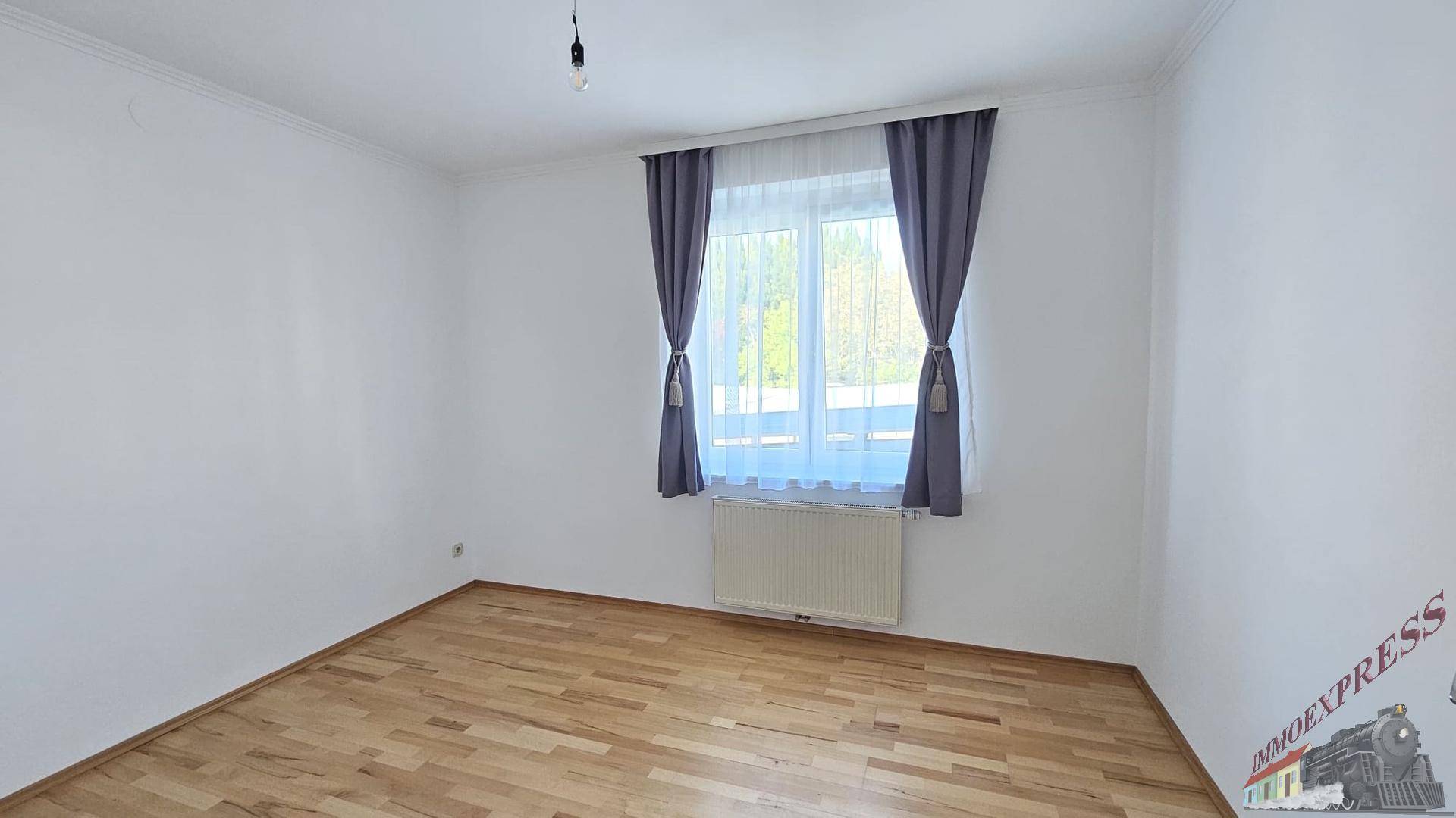 Schlafzimmer mit Parkettboden, weiß gestrichenen Wänden und einem Fenster mit Vorhängen.