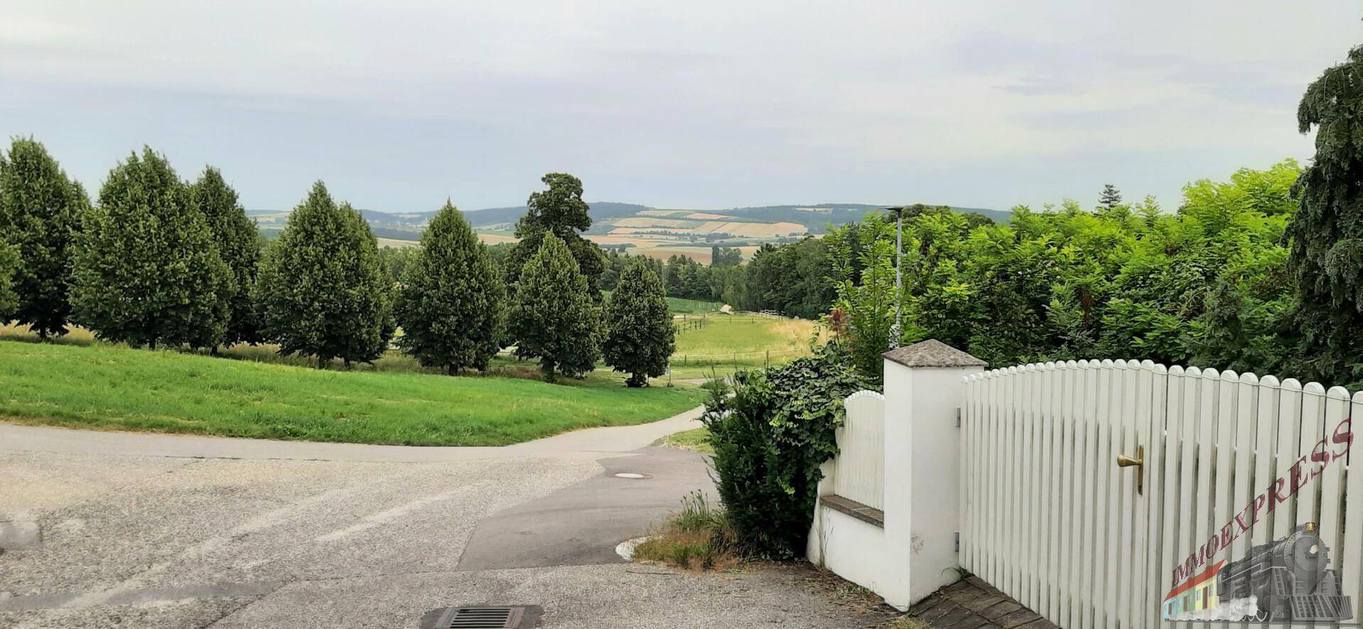 Zufahrtsstraße mit weißem Zaun und weitem Blick über grüne Hügel und Bäume.