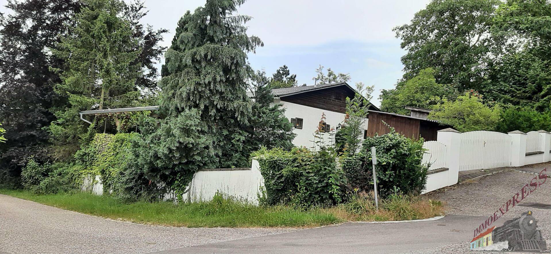 Hausansicht von der Straßenecke, umgeben von üppigem Grün und einem weißen Zaun.