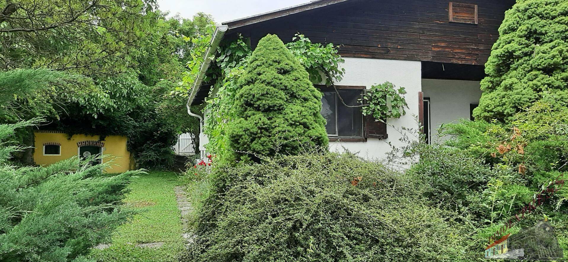 Außenansicht des Hauses mit üppigem Grün, Bäumen und einem kleinen Nebengebäude.