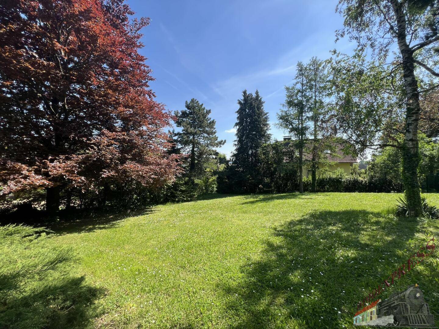 Großer, gepflegter Garten mit Rasenfläche und verschiedenen Bäumen unter strahlend blauem Himmel.