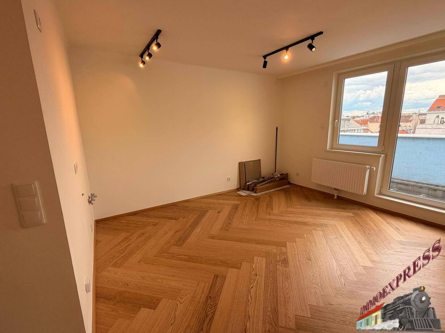 Lichtdurchfluteter Wohnraum mit großen Fenstern, Fischgrätparkett und Blick über die Dächer der Stadt.