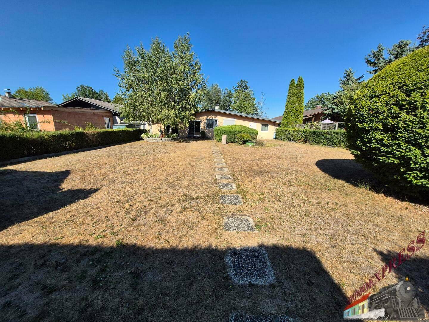Weitläufiger Gartenbereich hinter dem Haus mit trockenem Rasen und hohen Hecken.