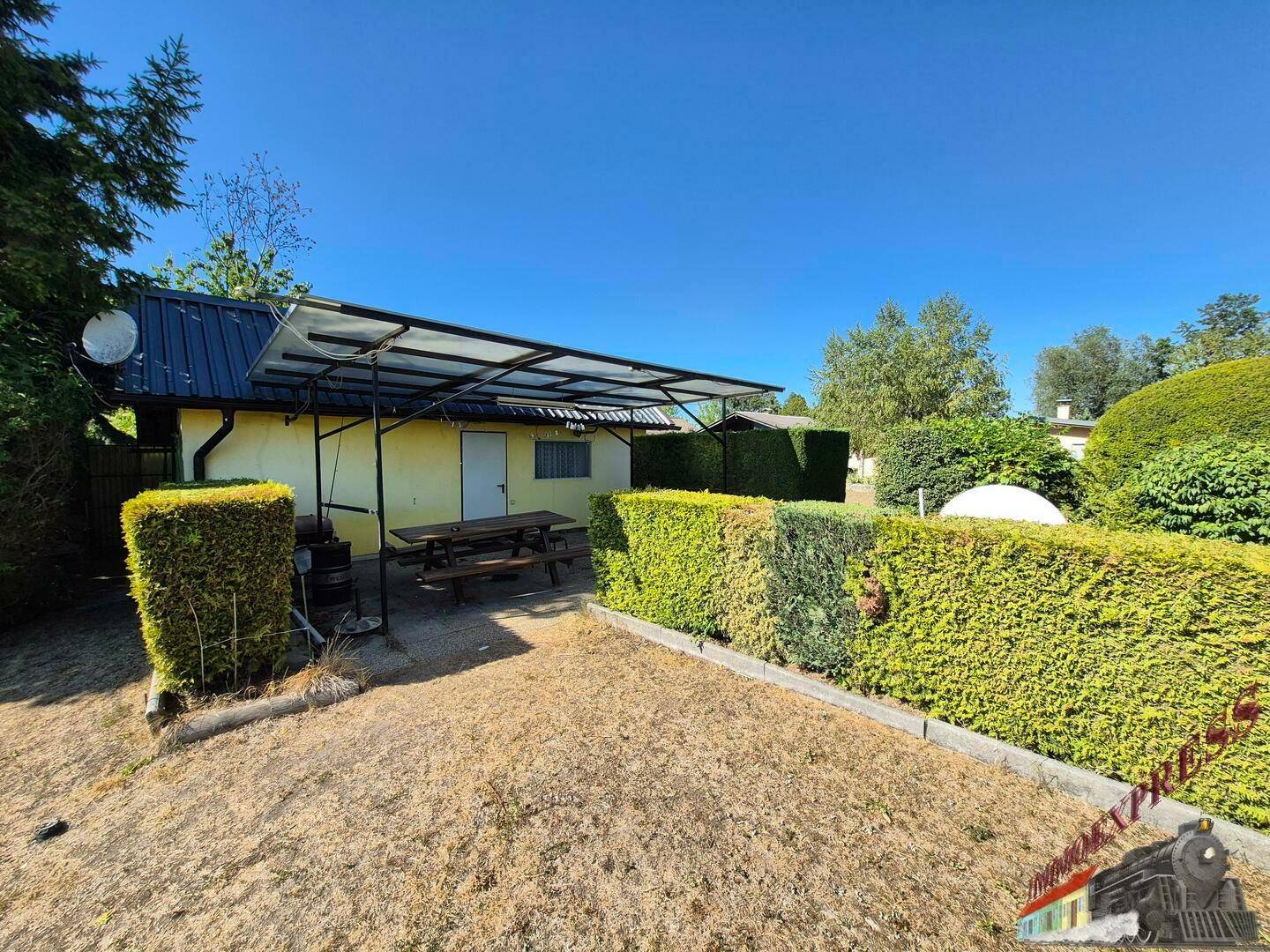 Außenansicht eines kleinen gelben Hauses mit überdachter Terrasse und gepflegten Hecken.