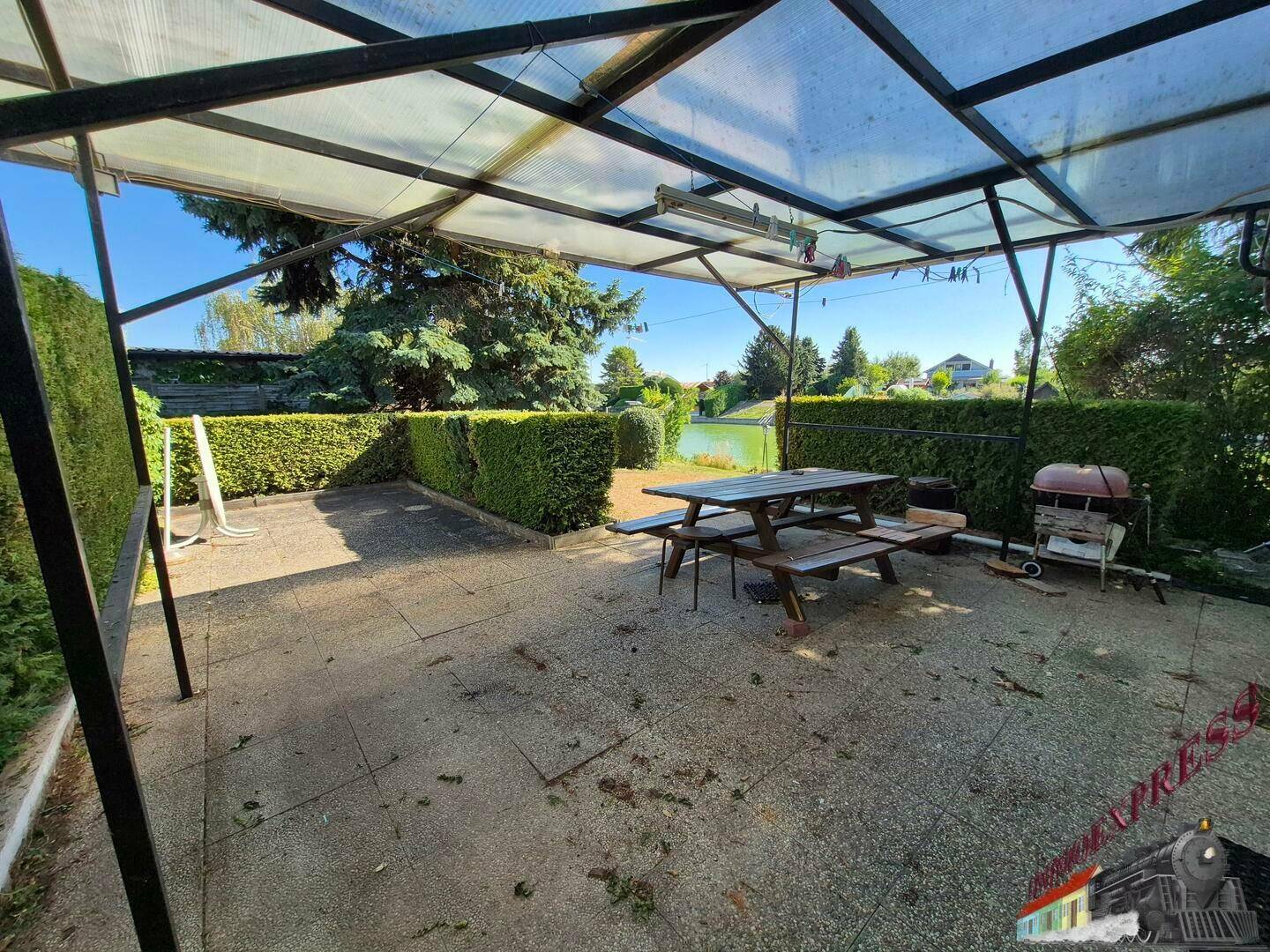 Überdachte Terrasse mit Picknicktisch und Grill, umgeben von grünen Hecken.