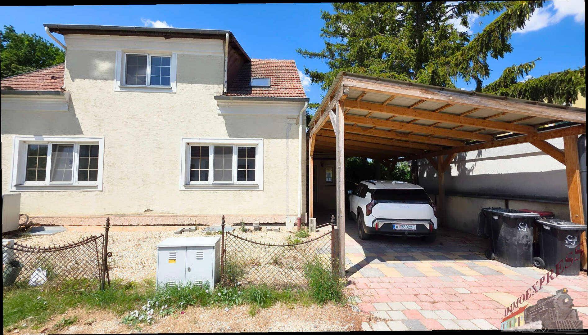 Freistehendes Haus mit heller Fassade und einem überdachten Carport auf gepflastertem Hof.