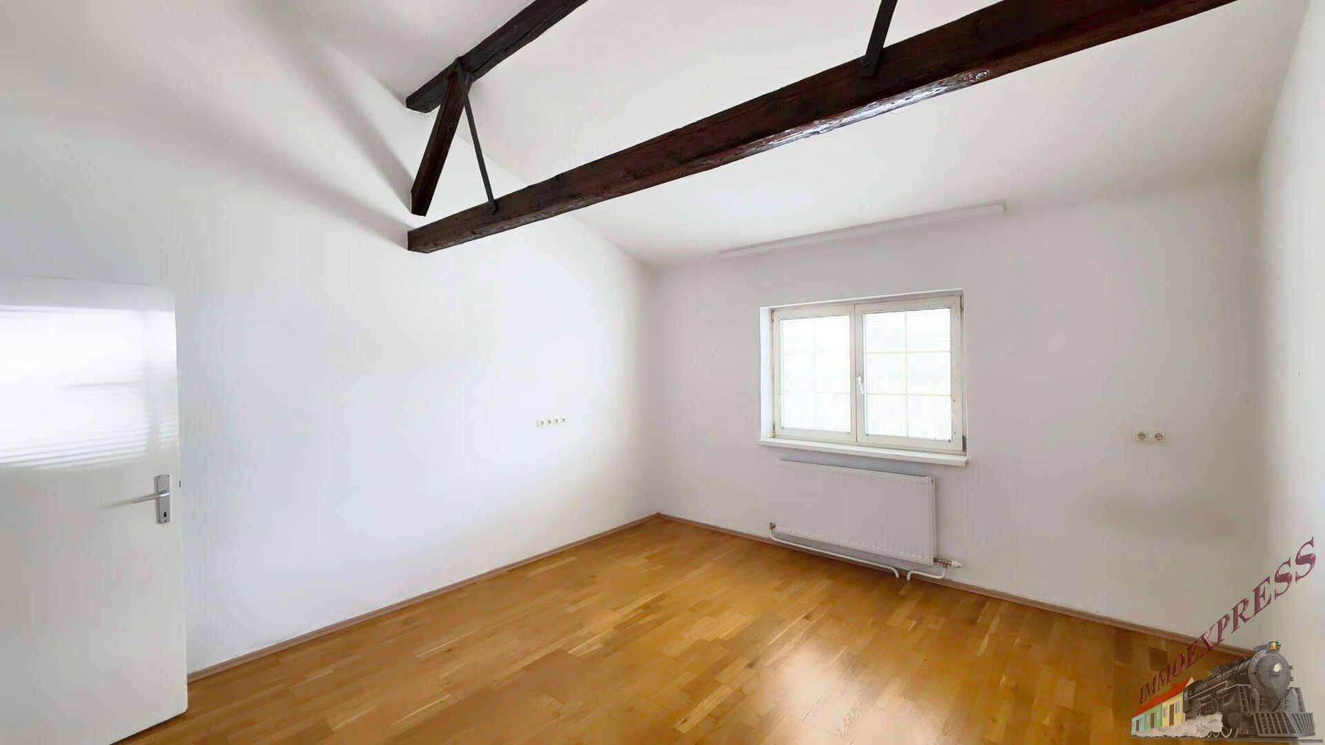 Helles Zimmer mit Holzboden, einem Fenster, weißen Wänden und sichtbaren Deckenbalken.