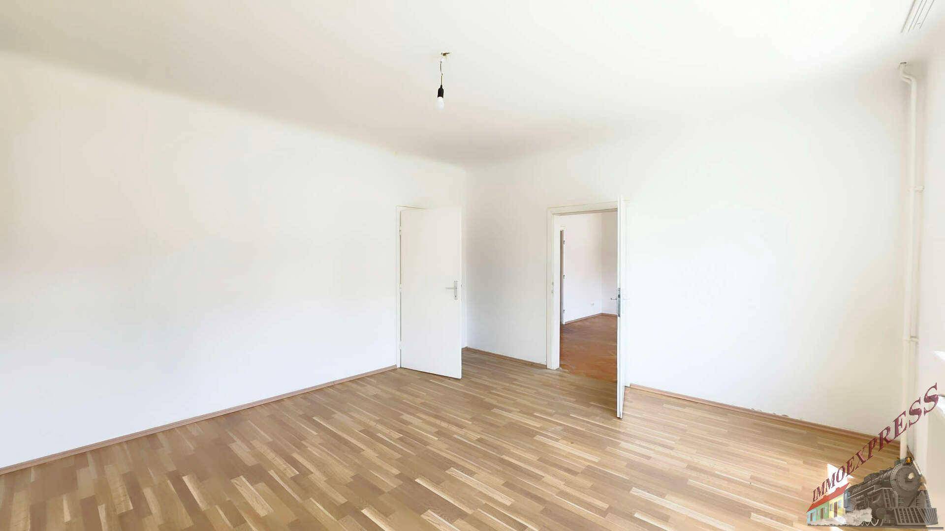 Geräumiges Zimmer mit Holzboden, weißen Wänden und zwei Türen, die zu weiteren Räumen führen.