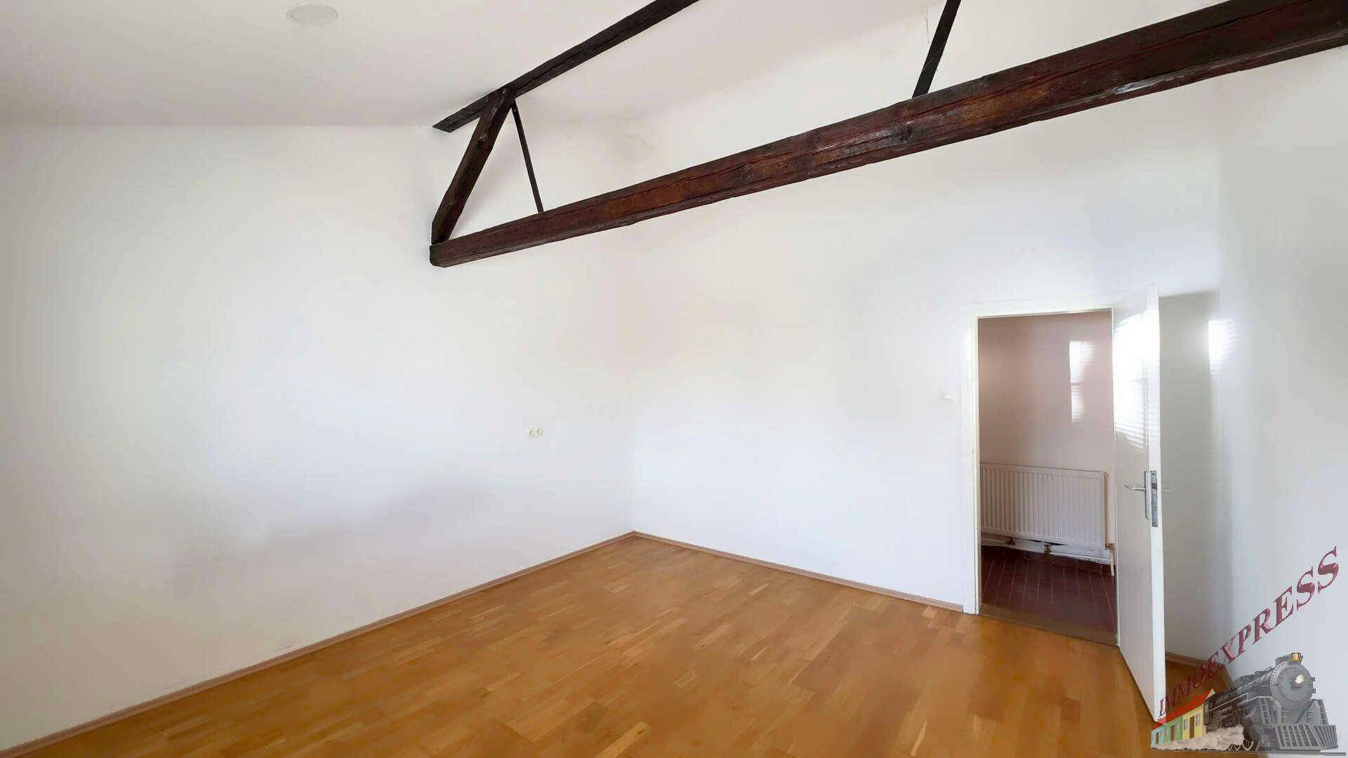 Helles Zimmer mit Holzboden, weißen Wänden und sichtbaren Holzbalken an der Decke.