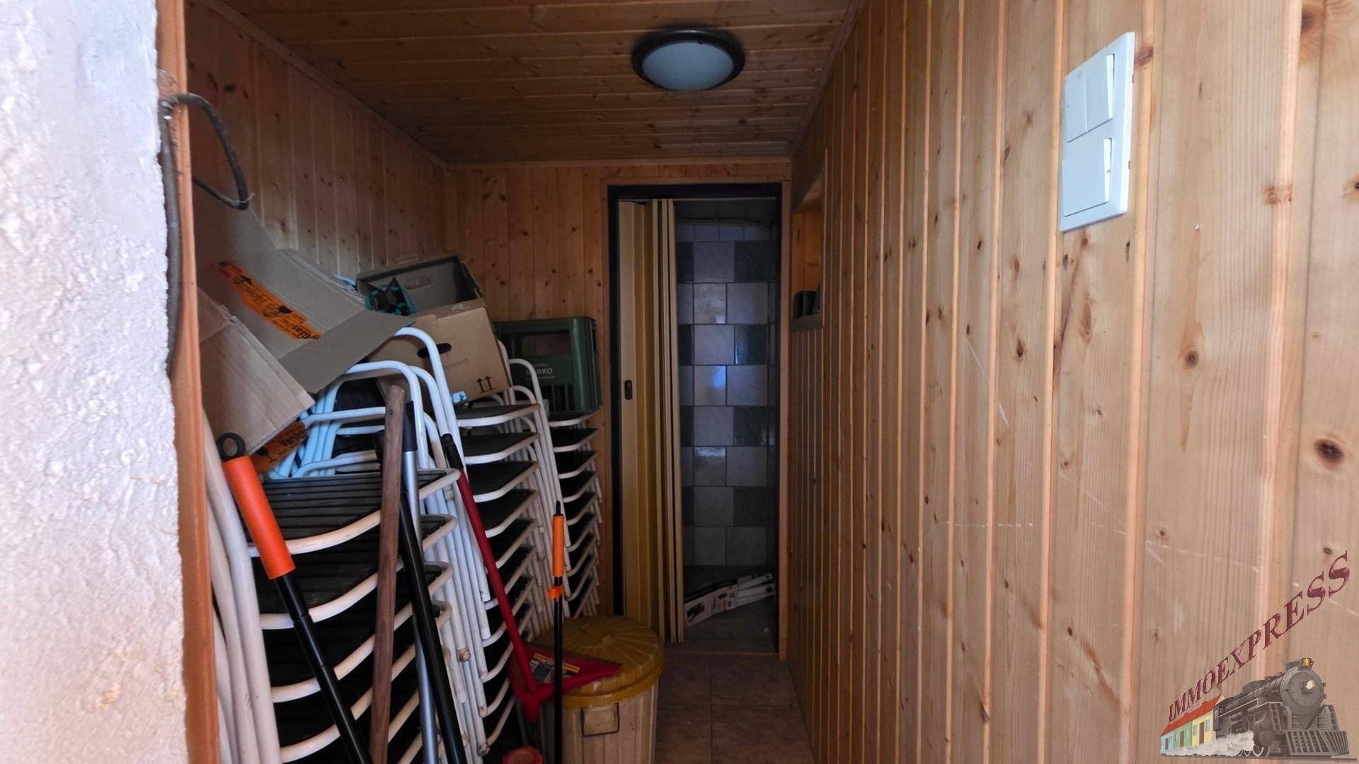 Schmaler Abstellraum mit Holzverkleidung und gestapelten Stühlen, führt zu einem gefliesten Bereich.