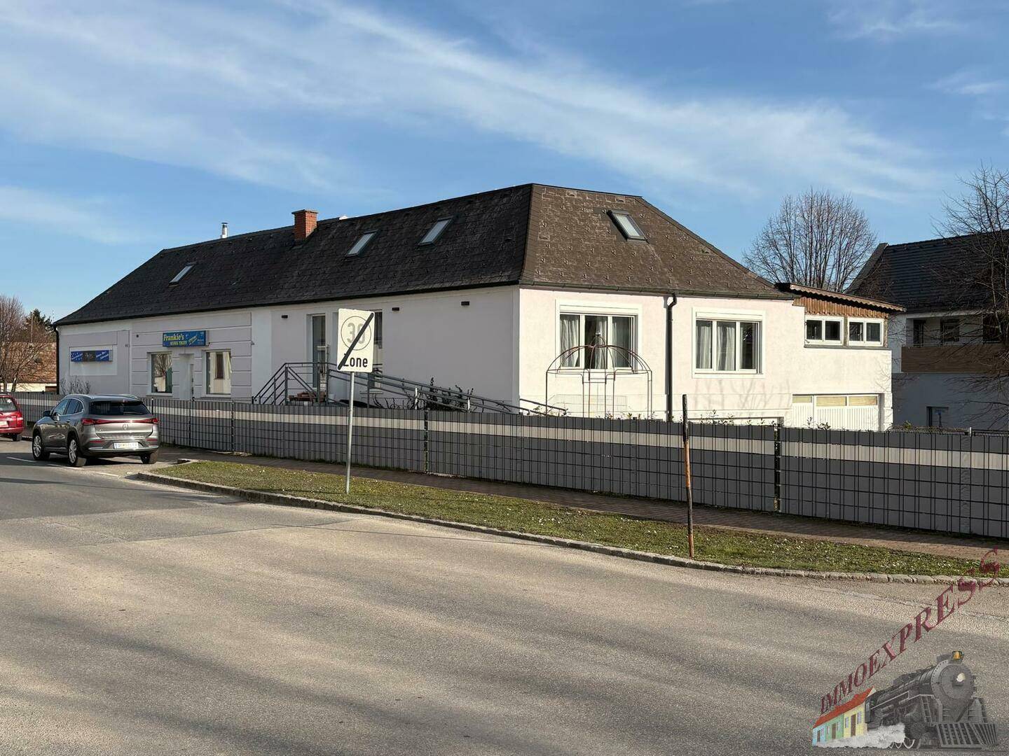 Gesamtansicht des Gebäudes von der Straße mit einem Zaun und parkenden Autos.