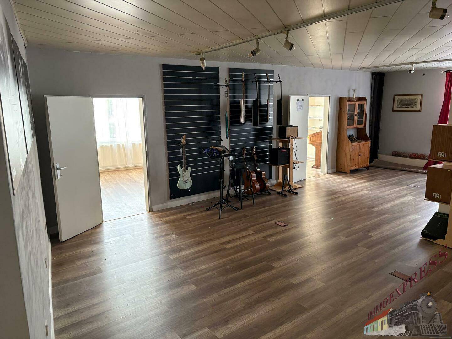 Großer Mehrzweckraum mit Holzboden, Musikinstrumenten und zwei Türen zu weiteren Zimmern.