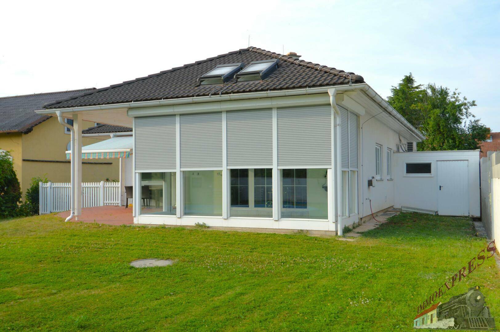 Seitenansicht des Hauses mit Wintergarten, Rollläden und gepflegter Rasenfläche im Vordergrund.