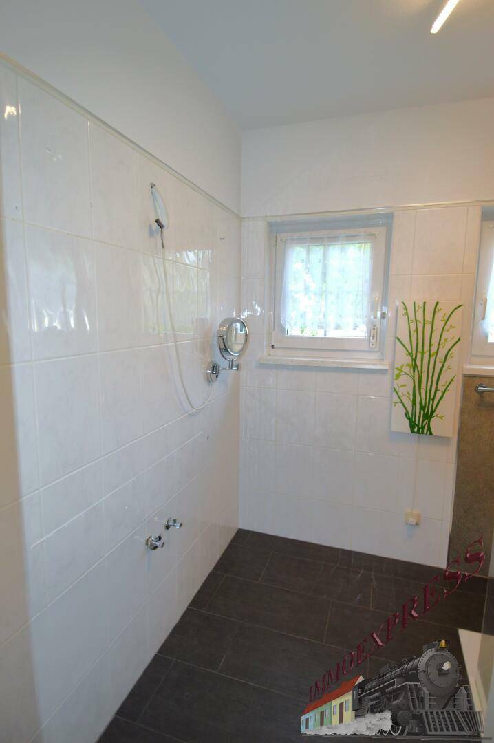 Helles Badezimmer mit weißen Wandfliesen, dunklem Boden und einer Duschvorrichtung.