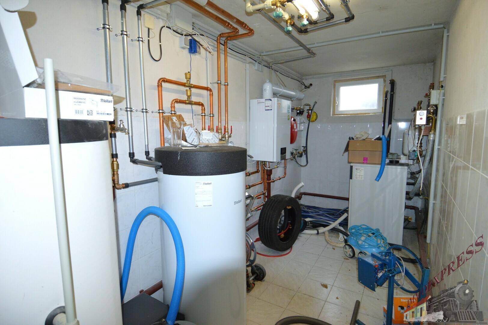 Technikraum mit Warmwasserspeichern, Rohrleitungen und kleinem Fenster.