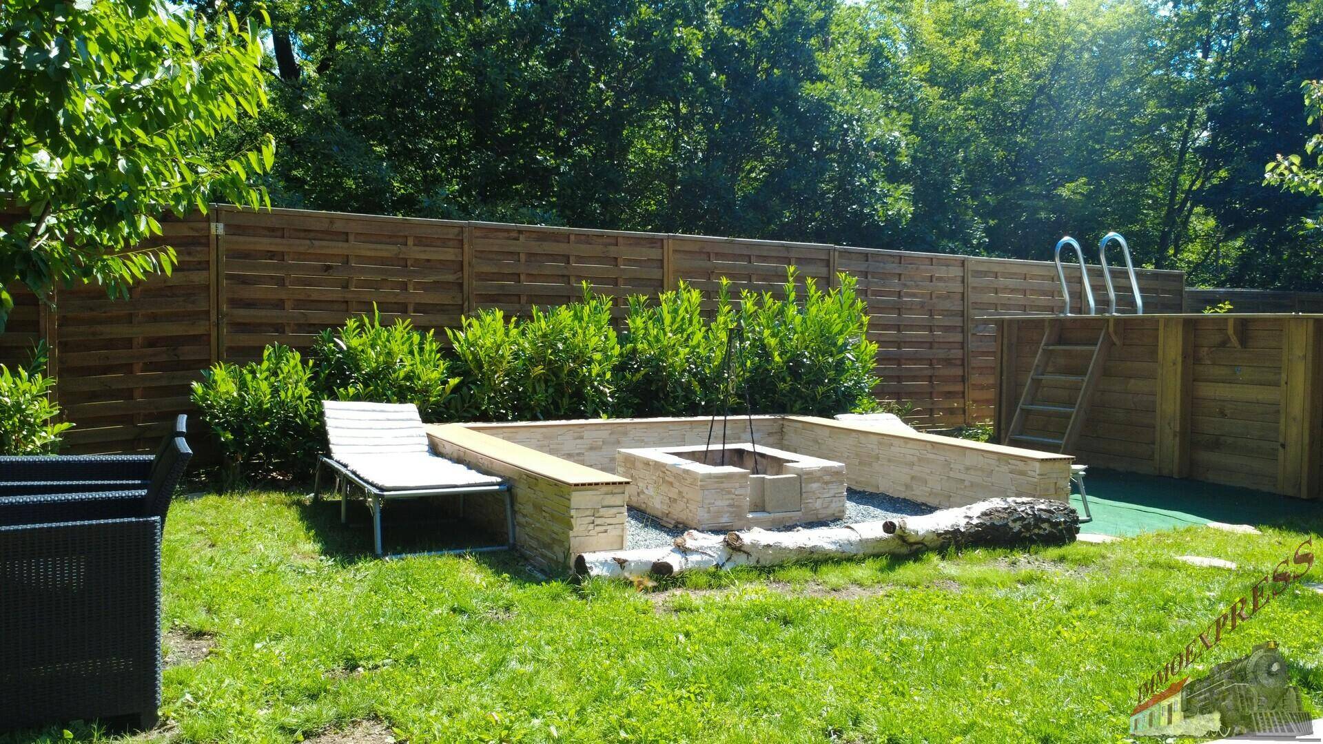 Gemütlicher Gartenbereich mit gemauerter Feuerstelle, Liegestühlen und erhöhtem Pool hinter einem Holzzaun.