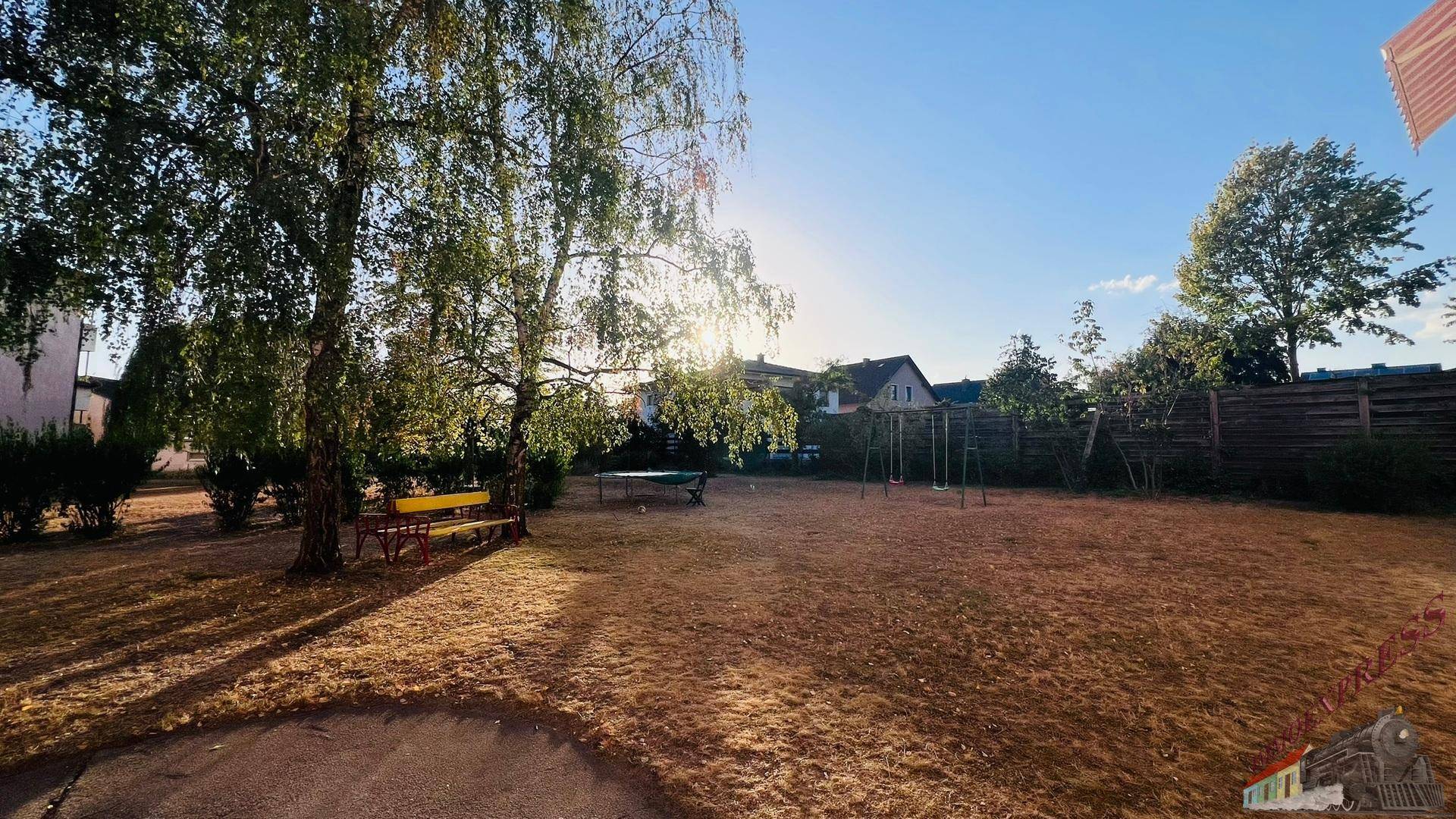 Großer Gartenbereich mit altem Baum, Sitzbank und Kinderschaukel unter strahlendem Sonnenschein.