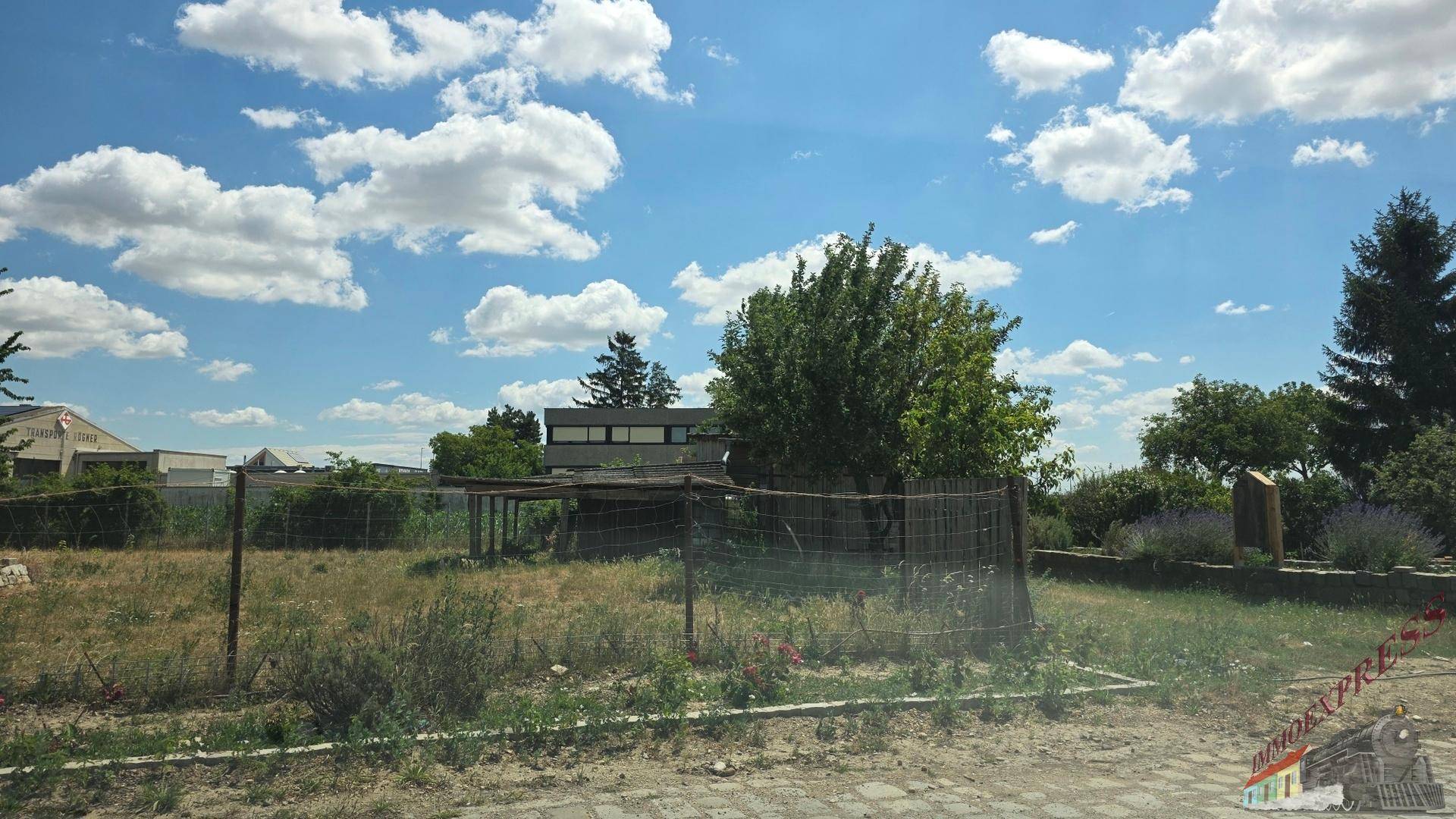 Ungenutztes Grundstück mit Zaun und Bäumen, im Hintergrund ein Gebäude unter blauem Himmel.