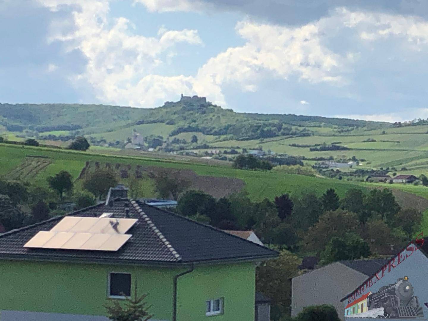Blick über Dächer mit Solaranlage auf grüne Hügel und ein historisches Schloss in der Ferne.