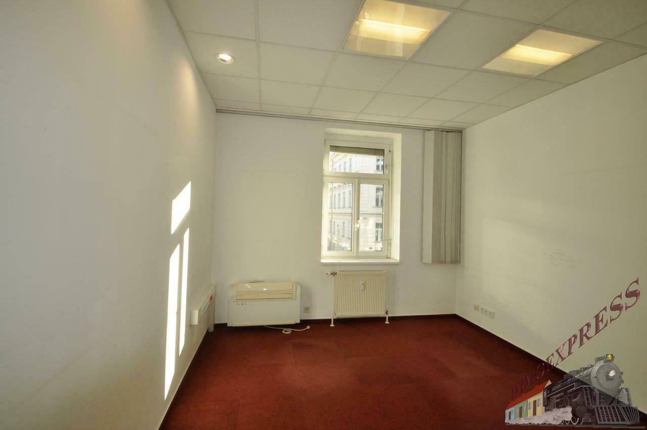 Helles Einzelbüro mit rotem Teppichboden, einem Fenster mit Jalousie und einem Heizkörper darunter.