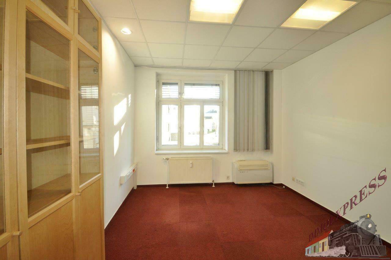 Bürozimmer mit rotem Teppichboden, einem Fenster, Heizkörper und einem hohen Holzschrank mit Glastüren.