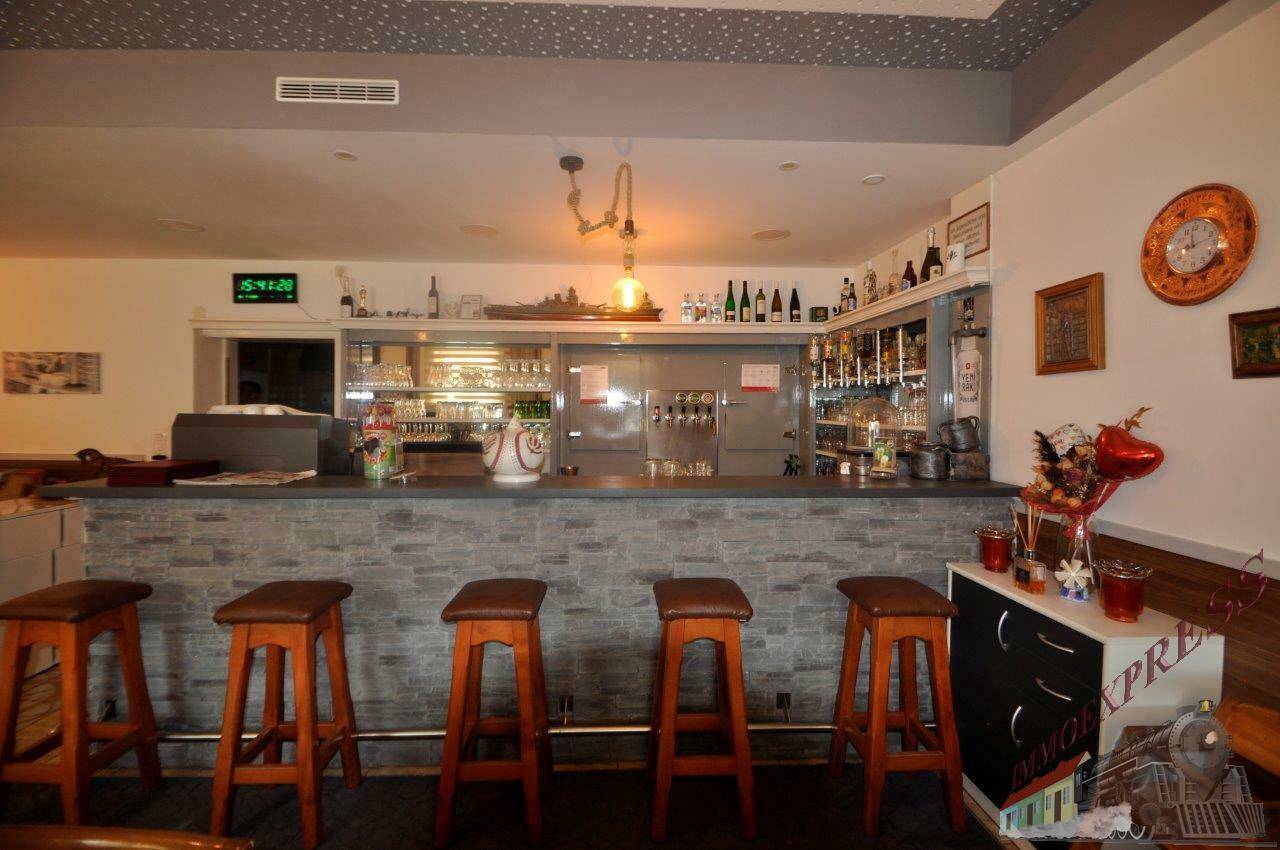 Innenansicht einer Bar mit Theke, Barhockern und Regalen voller Gläser und Flaschen.