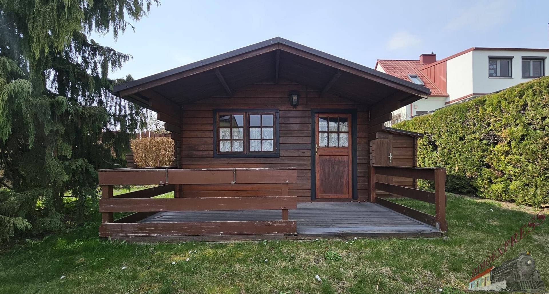 Kleine Holzhütte mit Veranda im Gartenbereich, umgeben von Grünflächen.