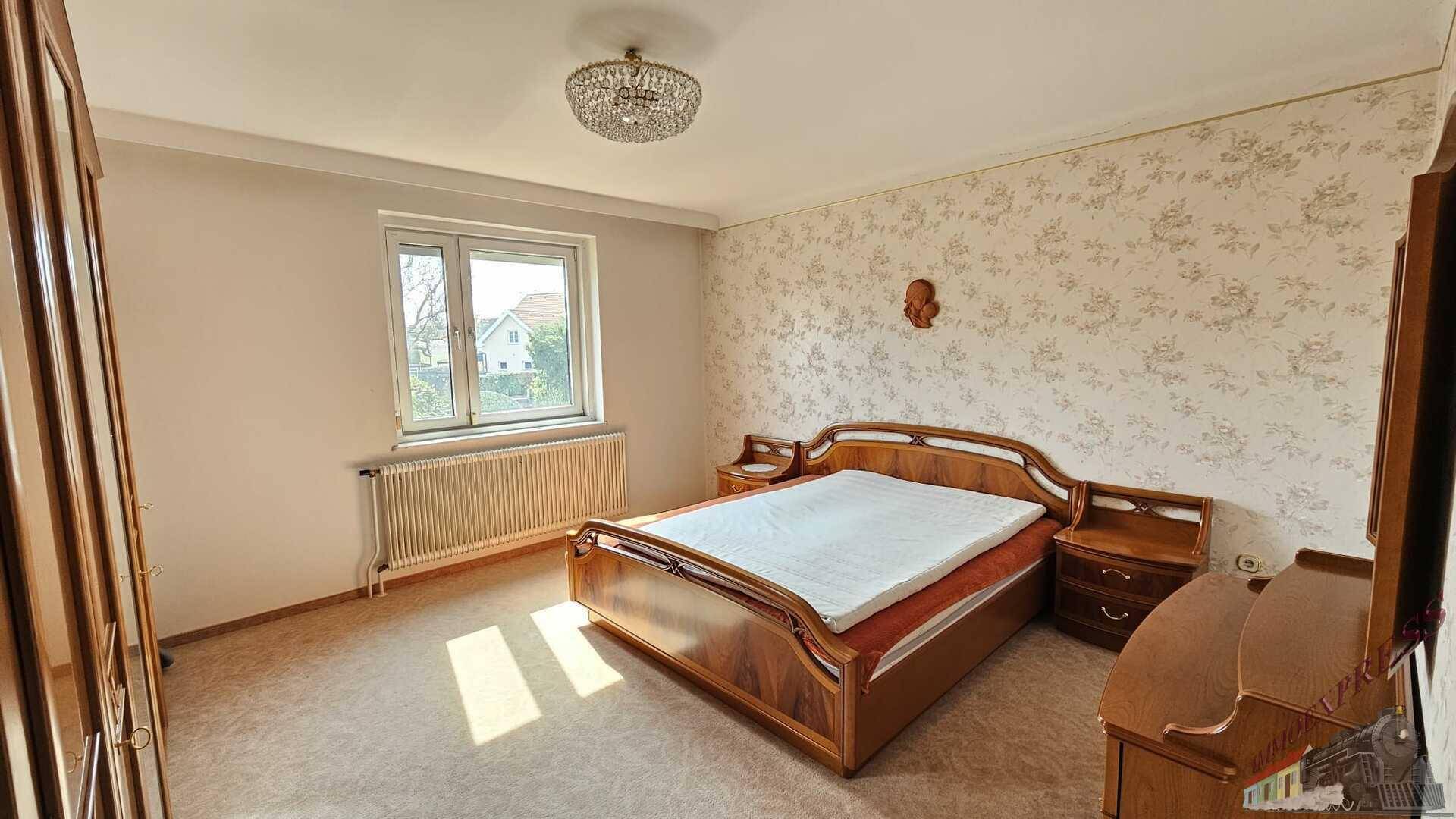 Schlafzimmer mit Doppelbett, floraler Tapete und Teppichboden, hell durch großes Fenster.
