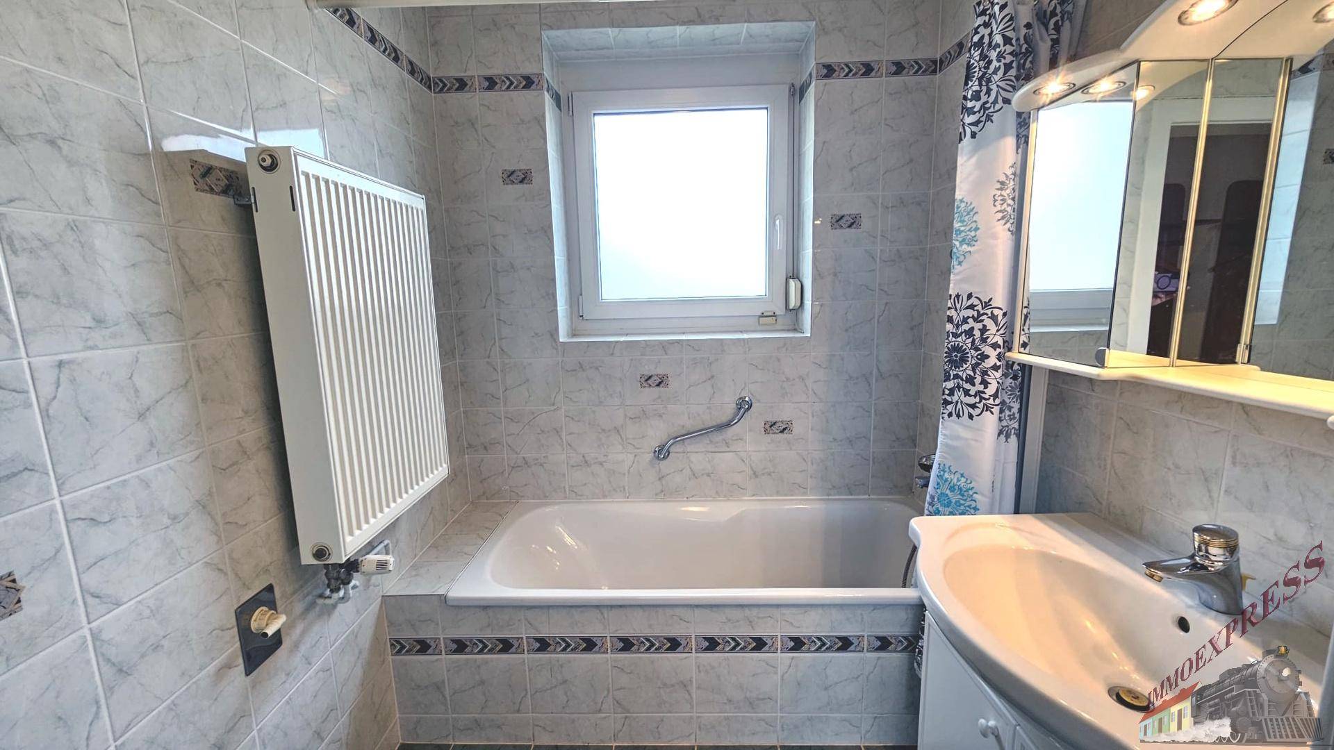 Badezimmer mit Badewanne, Fliesen und Fenster, inklusive Spiegelschrank und Einzelwaschbecken.